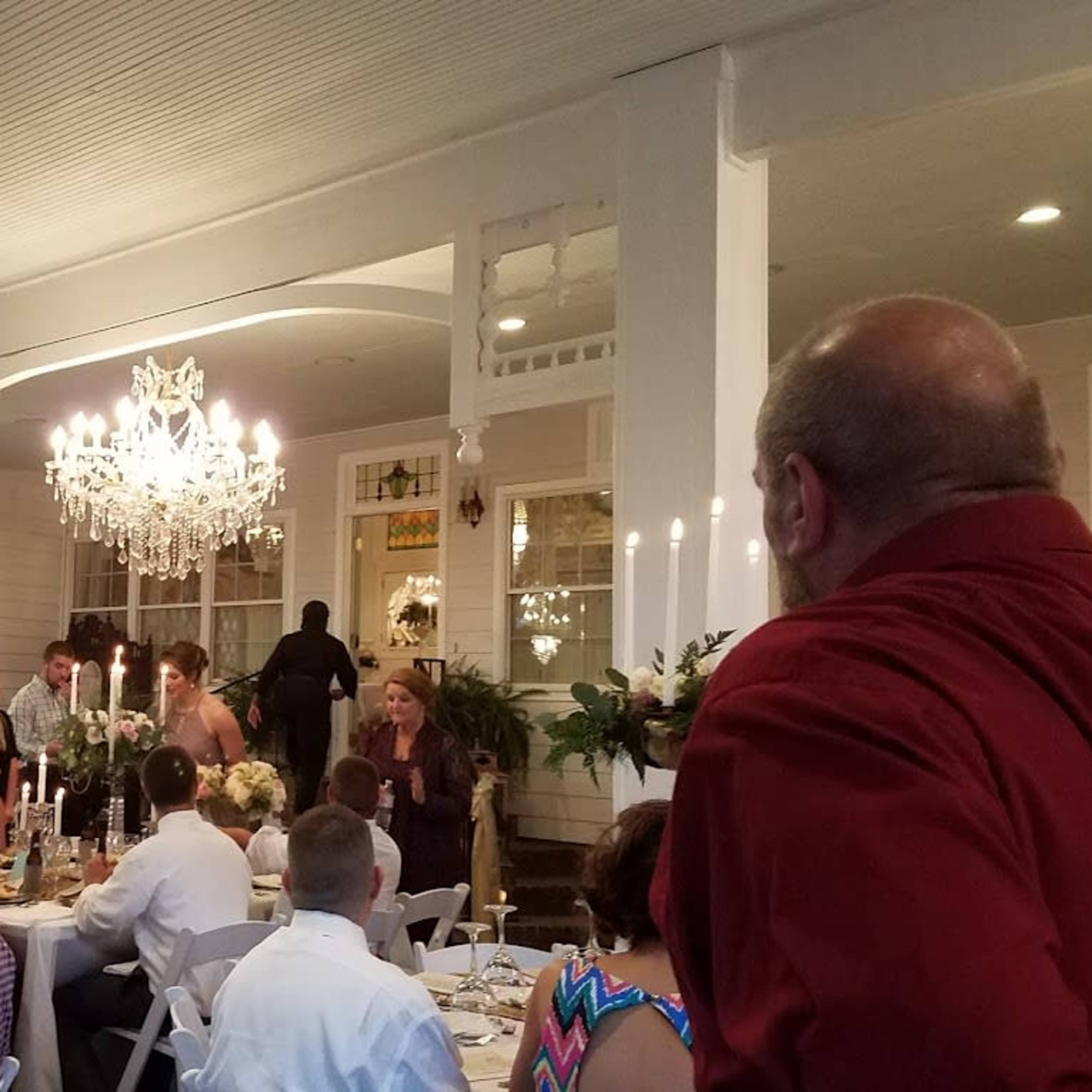 The scene depicts a dining event in a well-decorated interior with guests seated at tables and a chandelier hanging above.