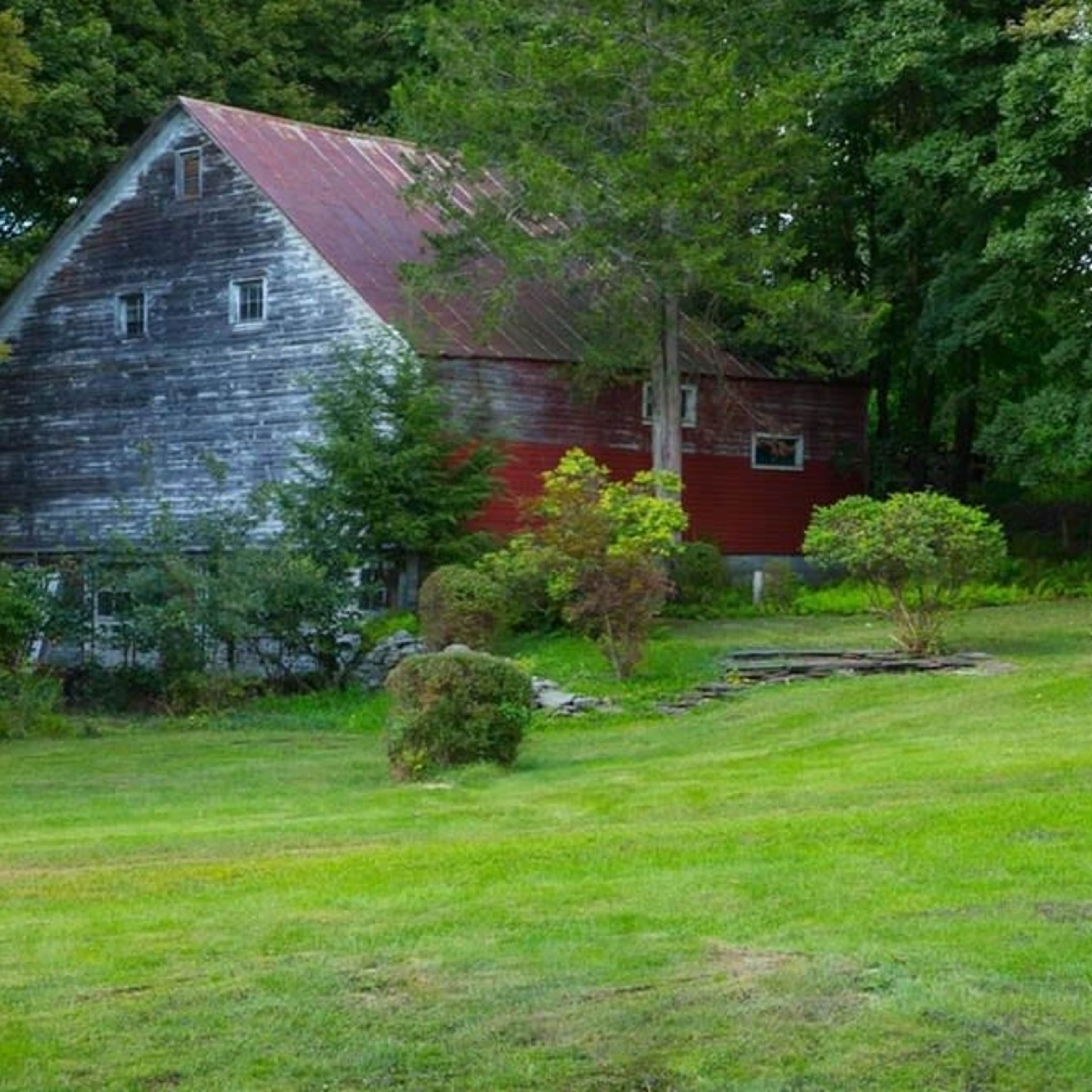 A weathered barn with a red section stands in a grassy area surrounded by trees and foliage.