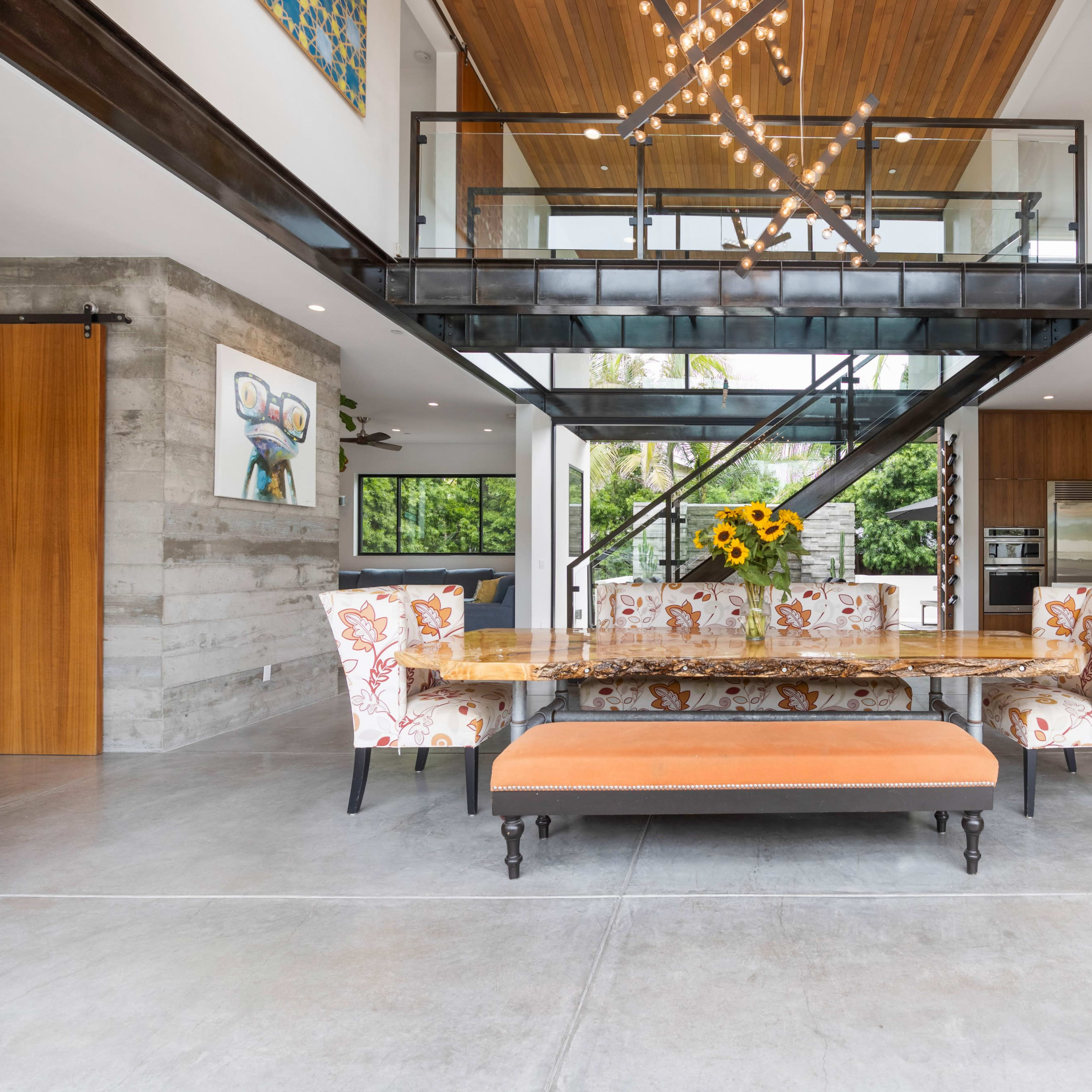 A modern, open-concept dining area featuring a large wooden table surrounded by floral-patterned chairs and a bench, under a ceiling with wooden beams and an artistic light fixture.