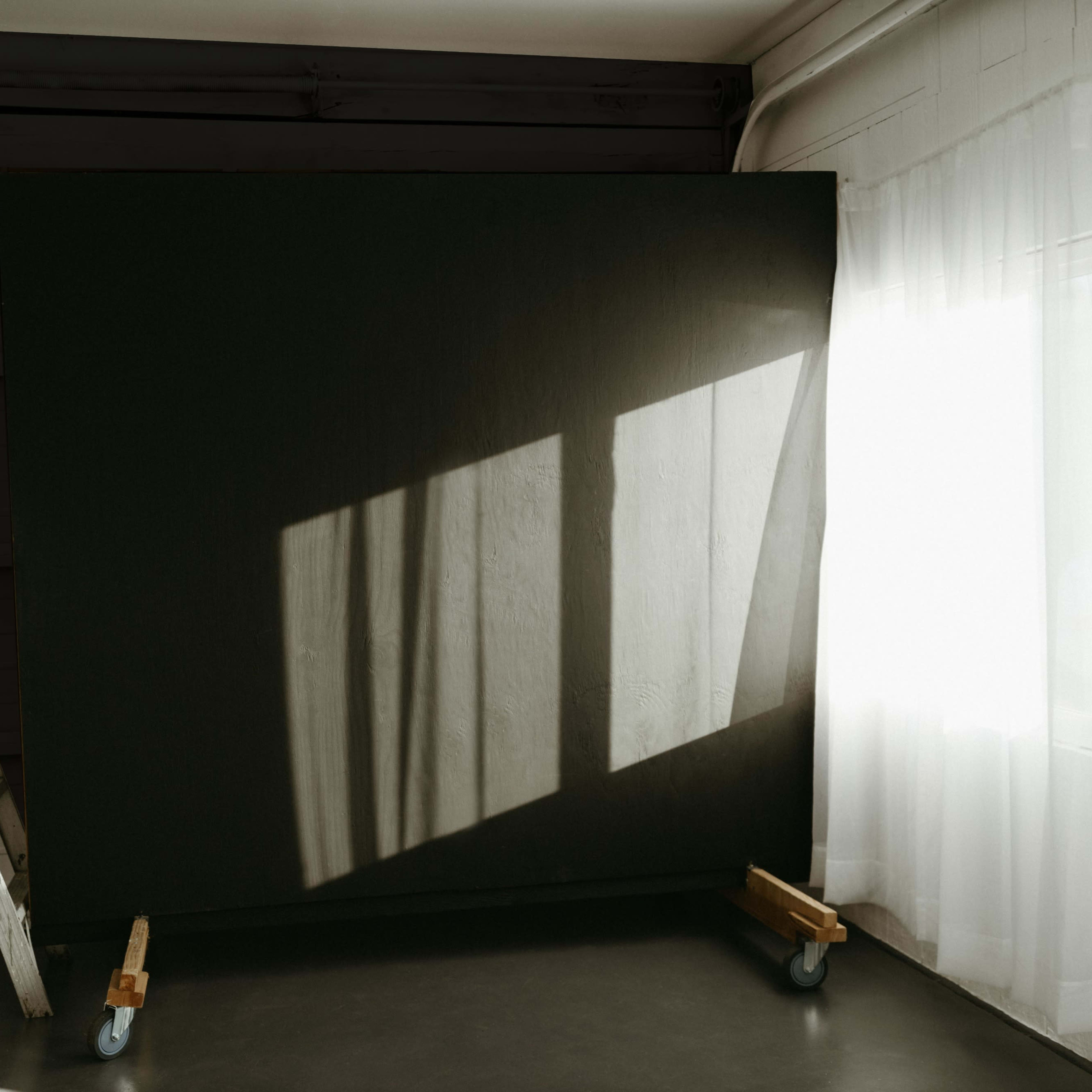 A large dark backdrop is positioned in a room with a ladder nearby, while sunlight casts shadows through a window onto the floor.