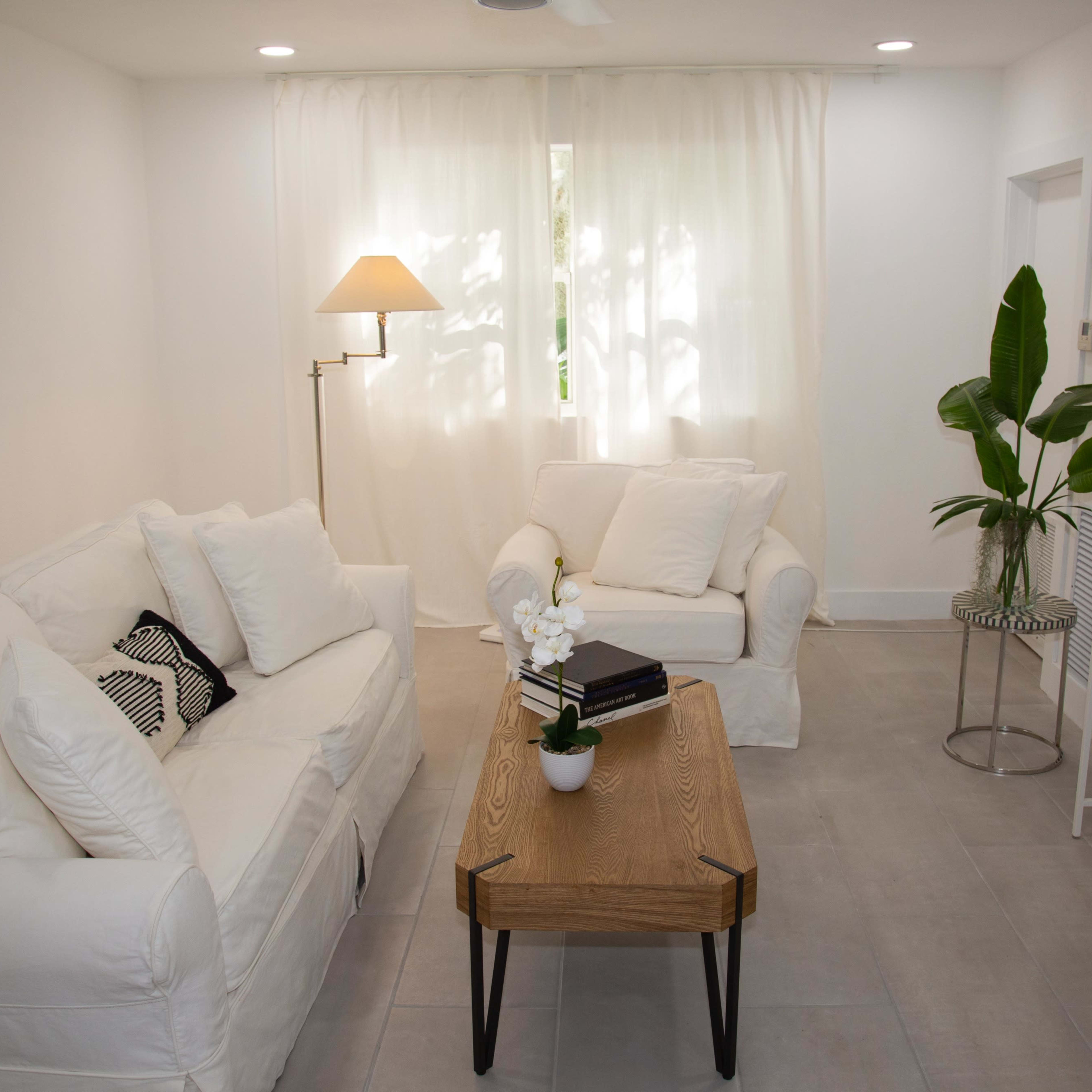 A bright living room features two white sofas, a wooden coffee table, a plant, and a television.