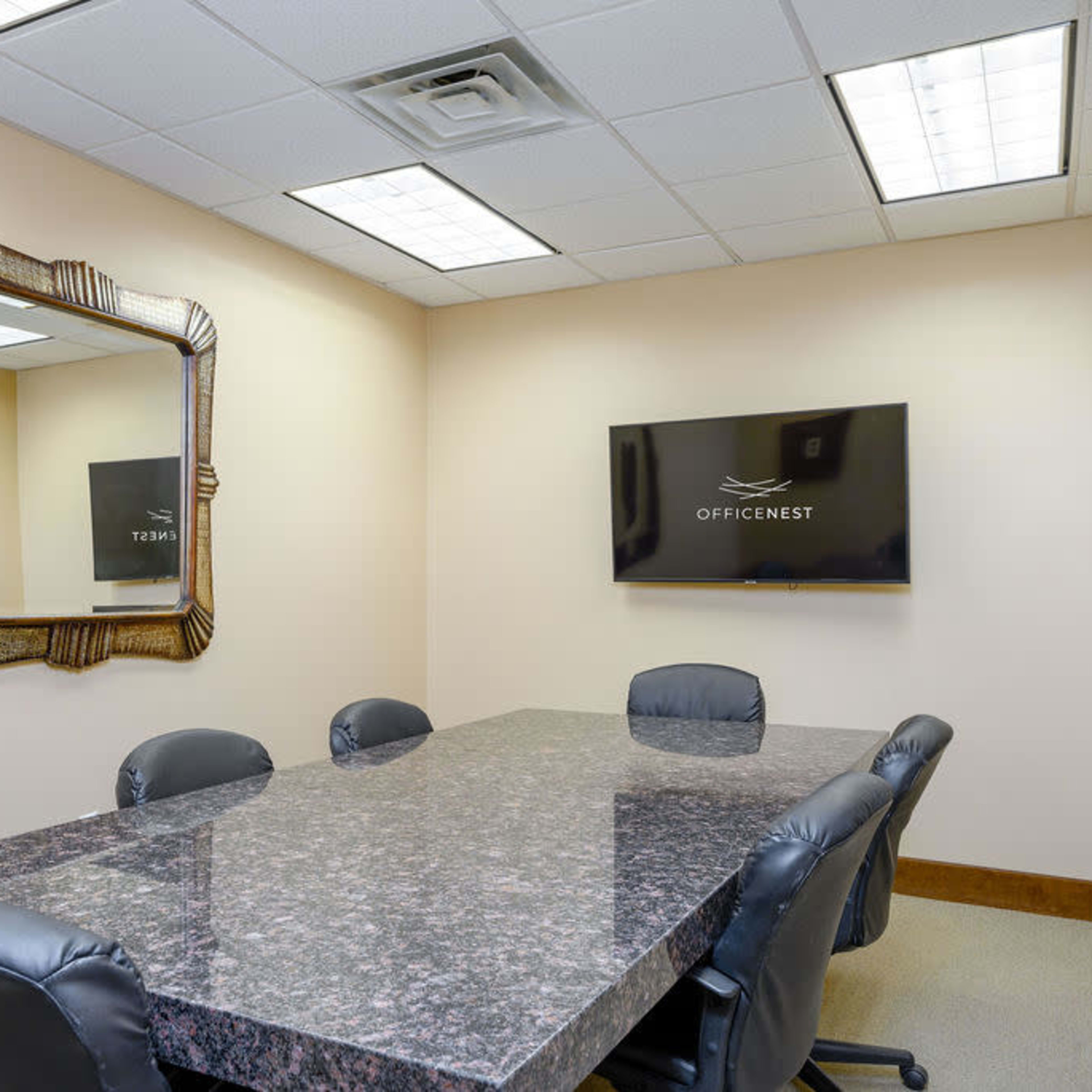 A small meeting room featuring a rectangular table surrounded by black leather chairs, a decorative mirror on the wall, and a wall-mounted TV displaying the logo "OFFICENEST."