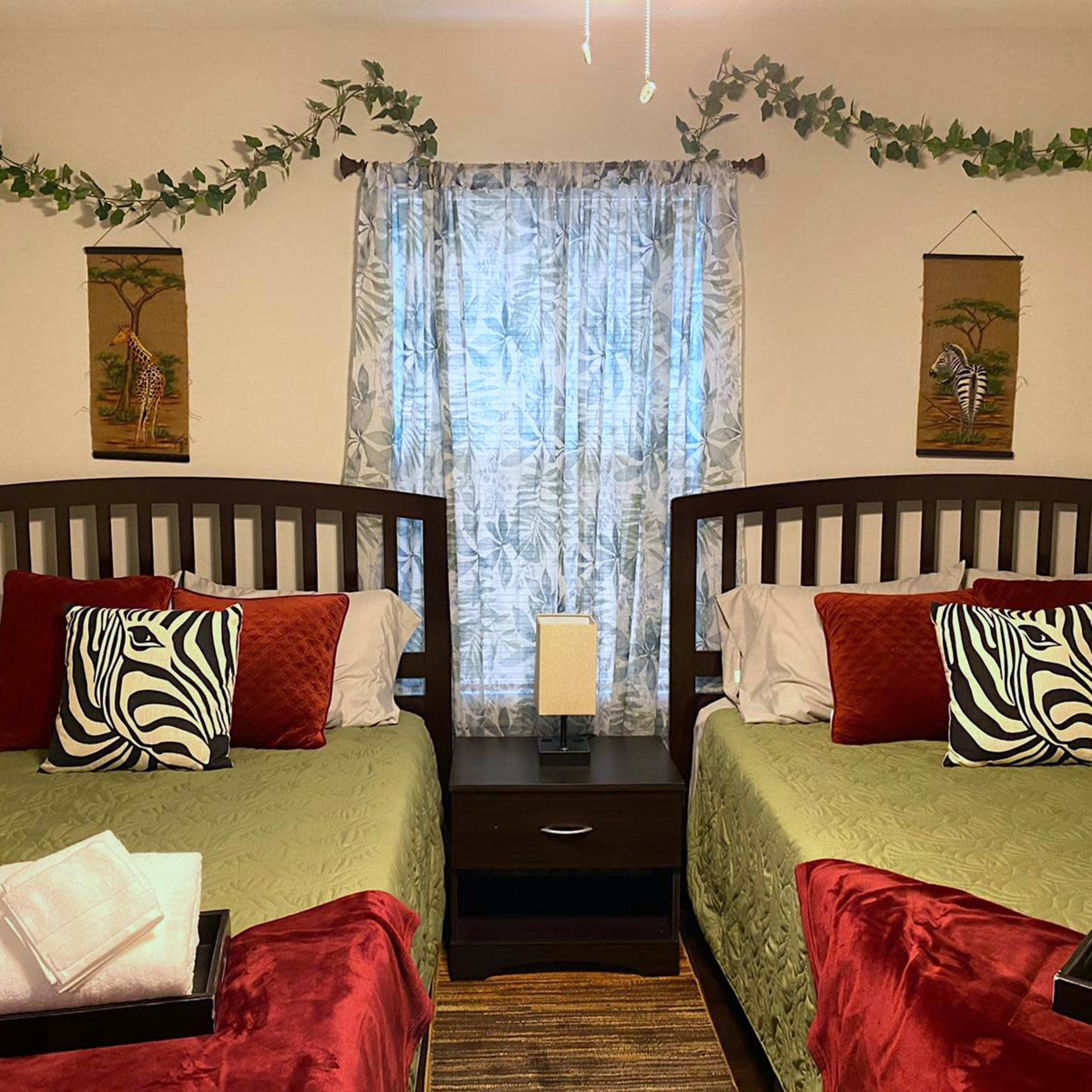 A cozy bedroom features two beds with red and green bedding, decorative zebra-print pillows, and artwork on the walls, set against a window dressed in sheer curtains.