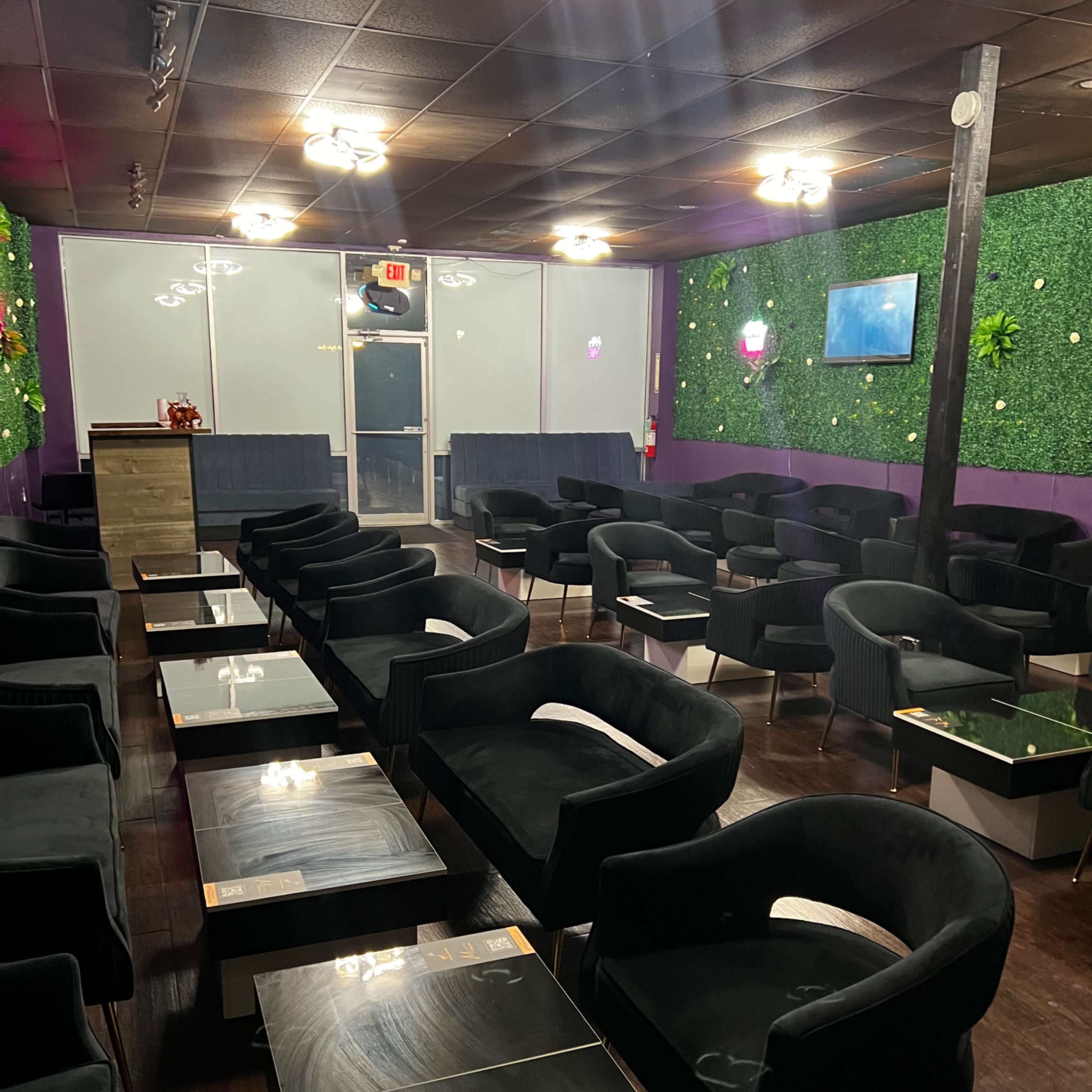 A lounge area with several black chairs and tables arranged in rows, surrounded by green wall decor and dim lighting.