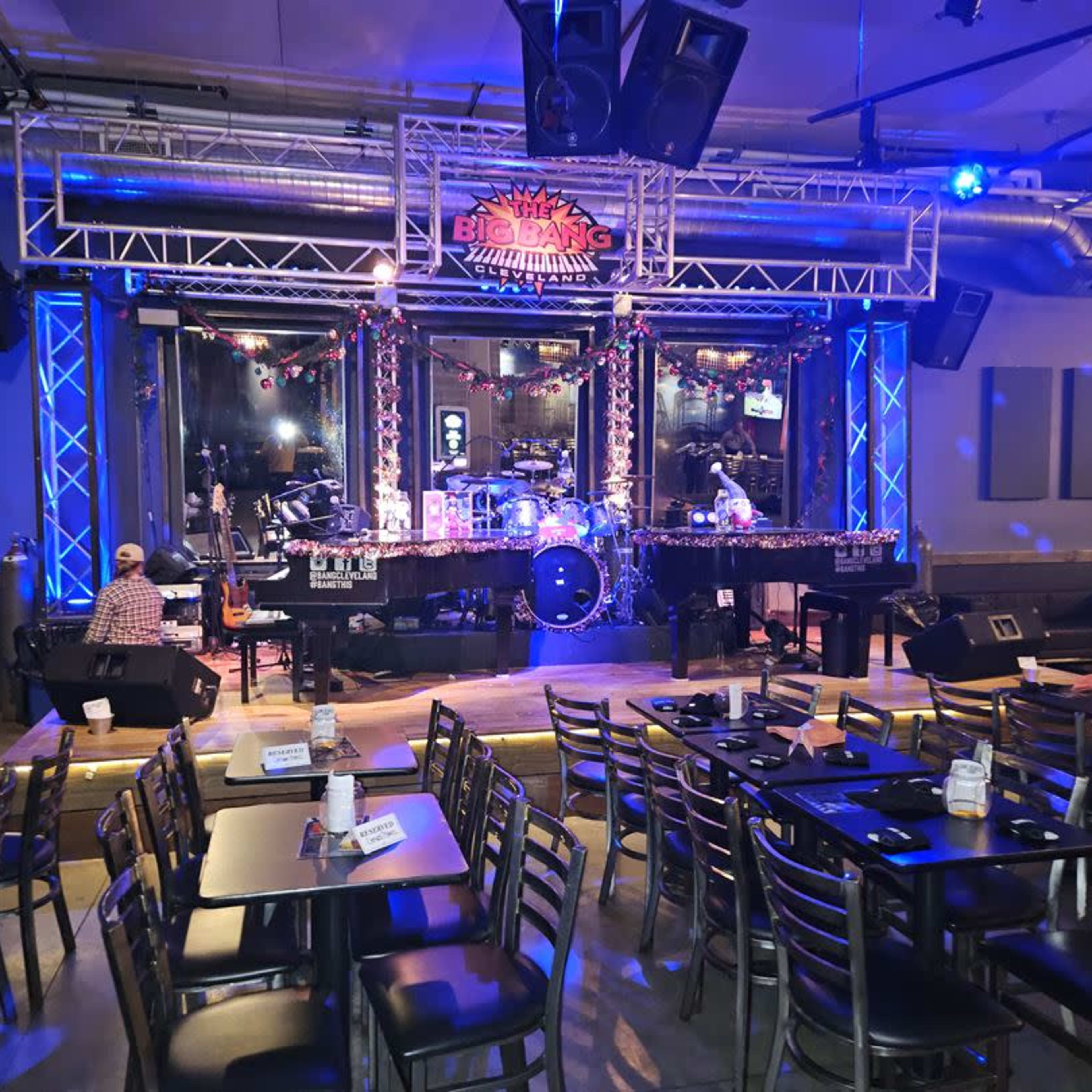 A live music venue features a stage with instruments set up under bright lights, surrounded by tables and chairs.
