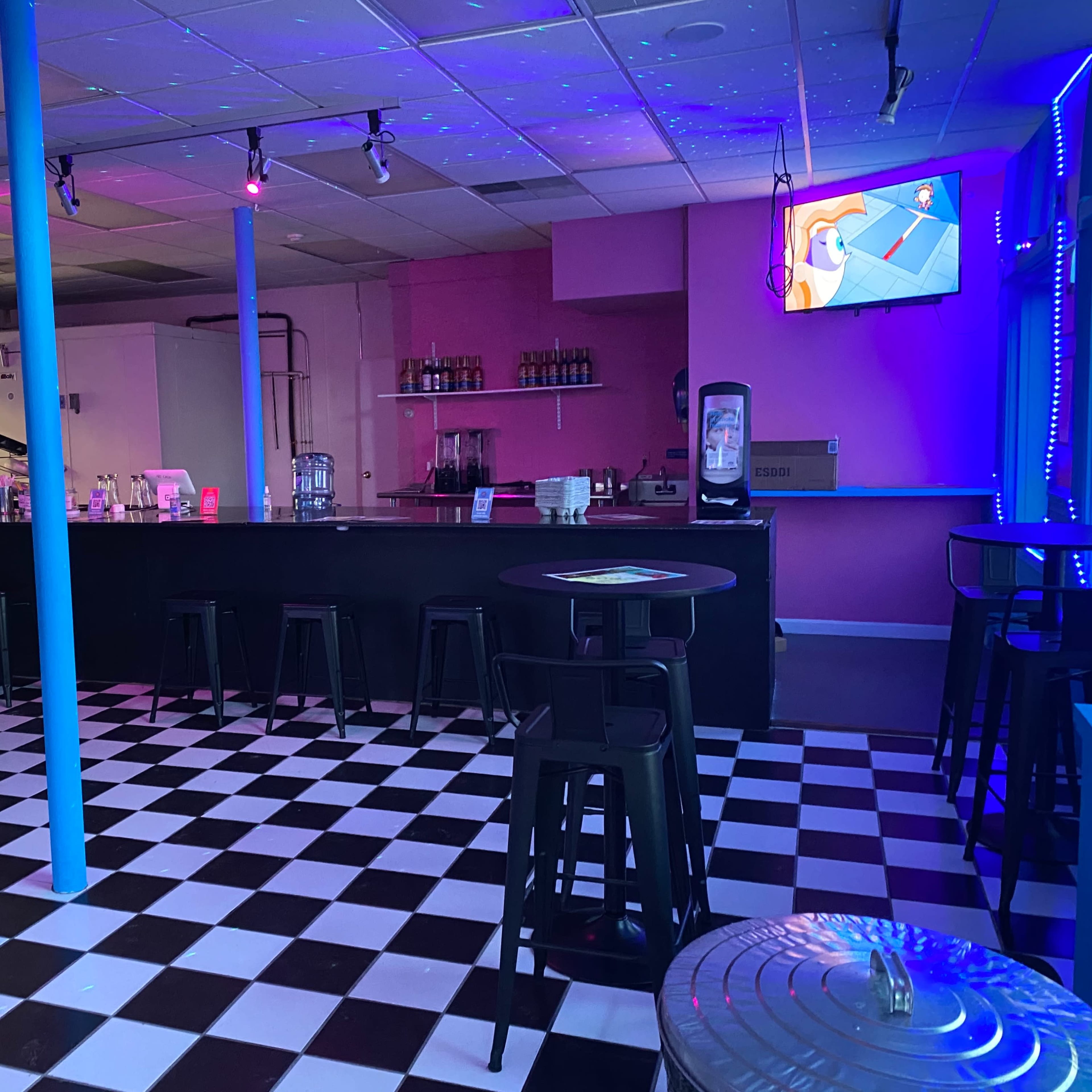 A colorful bar area with a checkered black and white floor, a pink wall, and a TV mounted on the right displaying a vibrant scene.