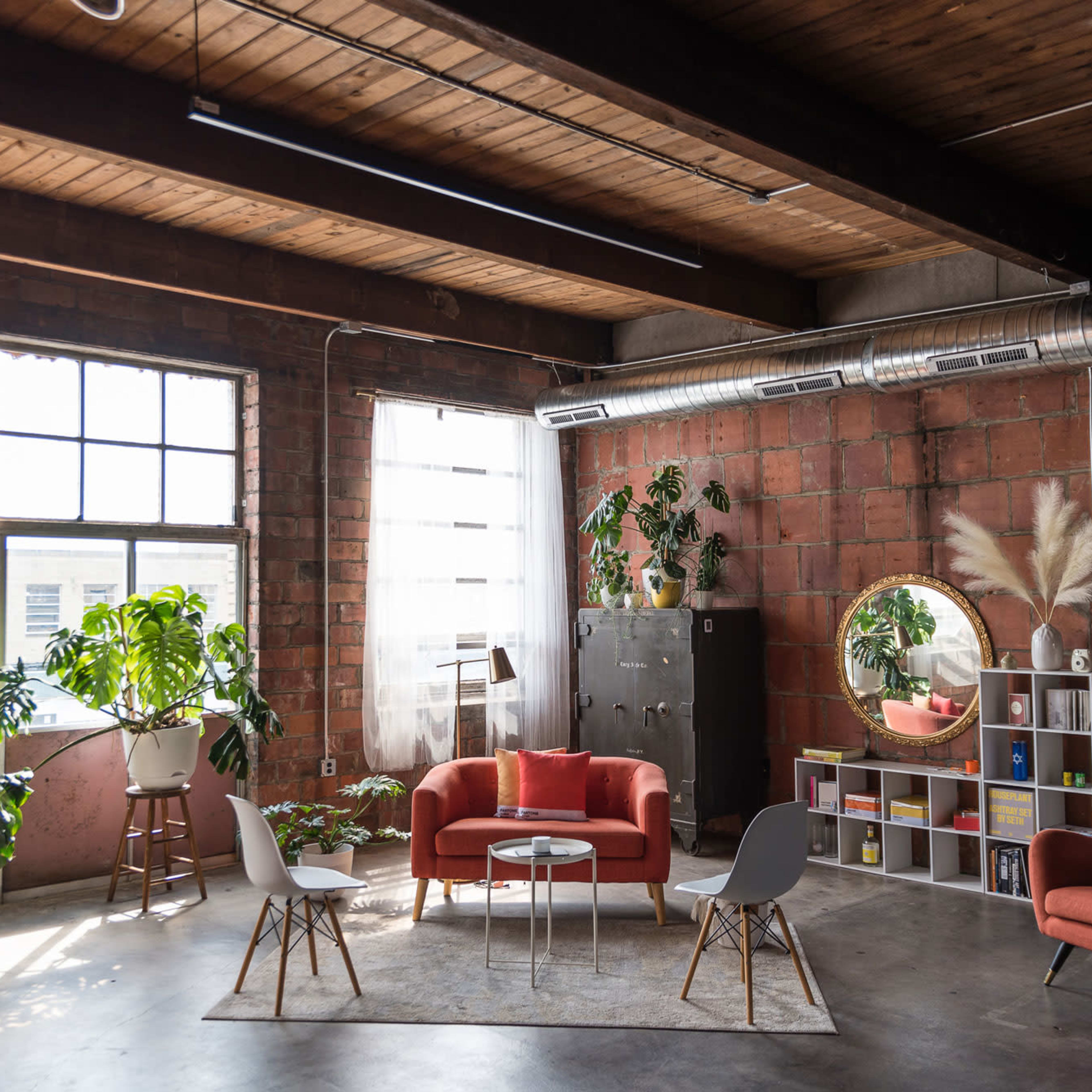 A modern, industrial-style living space with exposed brick walls, large windows, and contemporary furniture arrangements including a red sofa and white chairs.