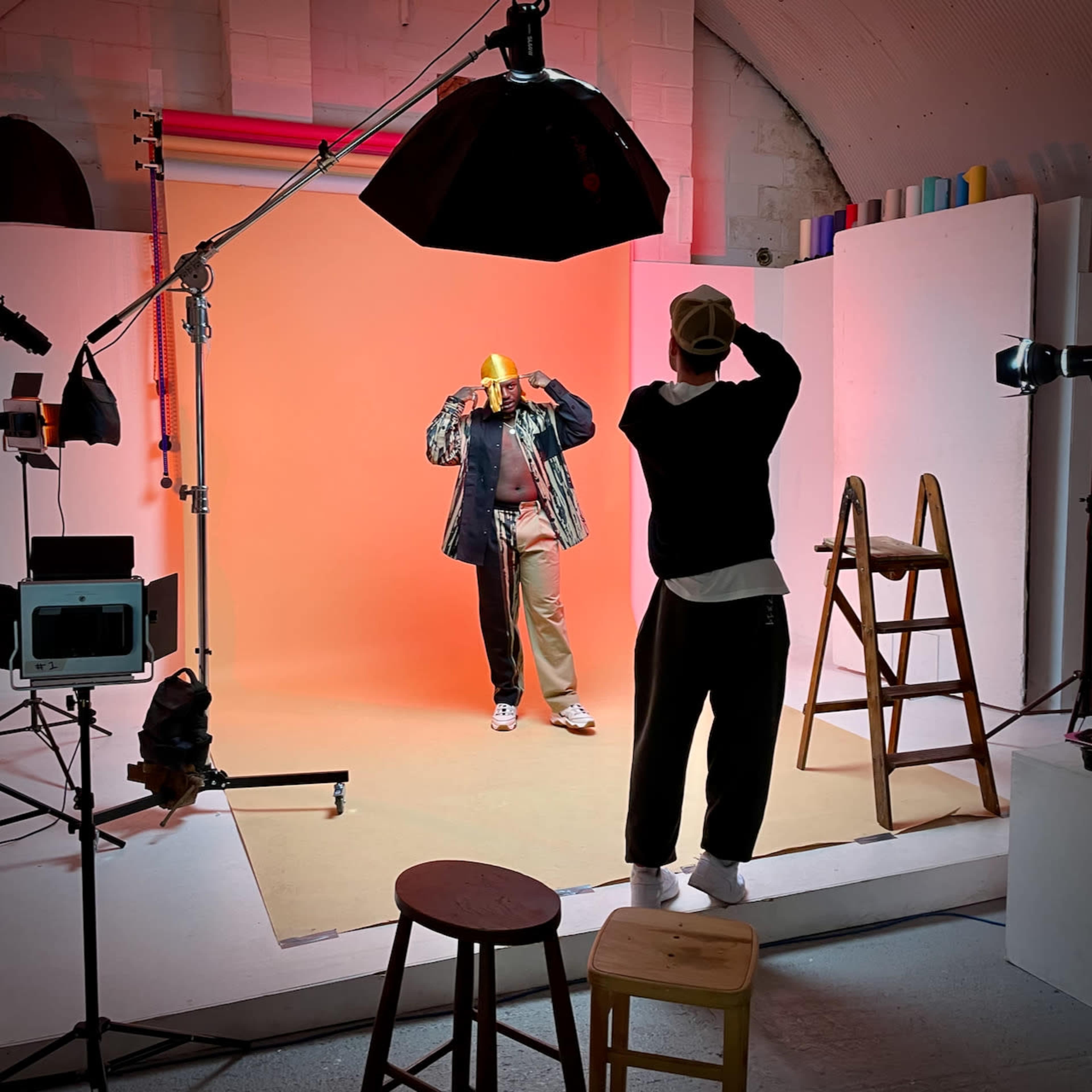 A person wearing a yellow hat poses in a studio with colorful backdrops while another person stands nearby, observing.