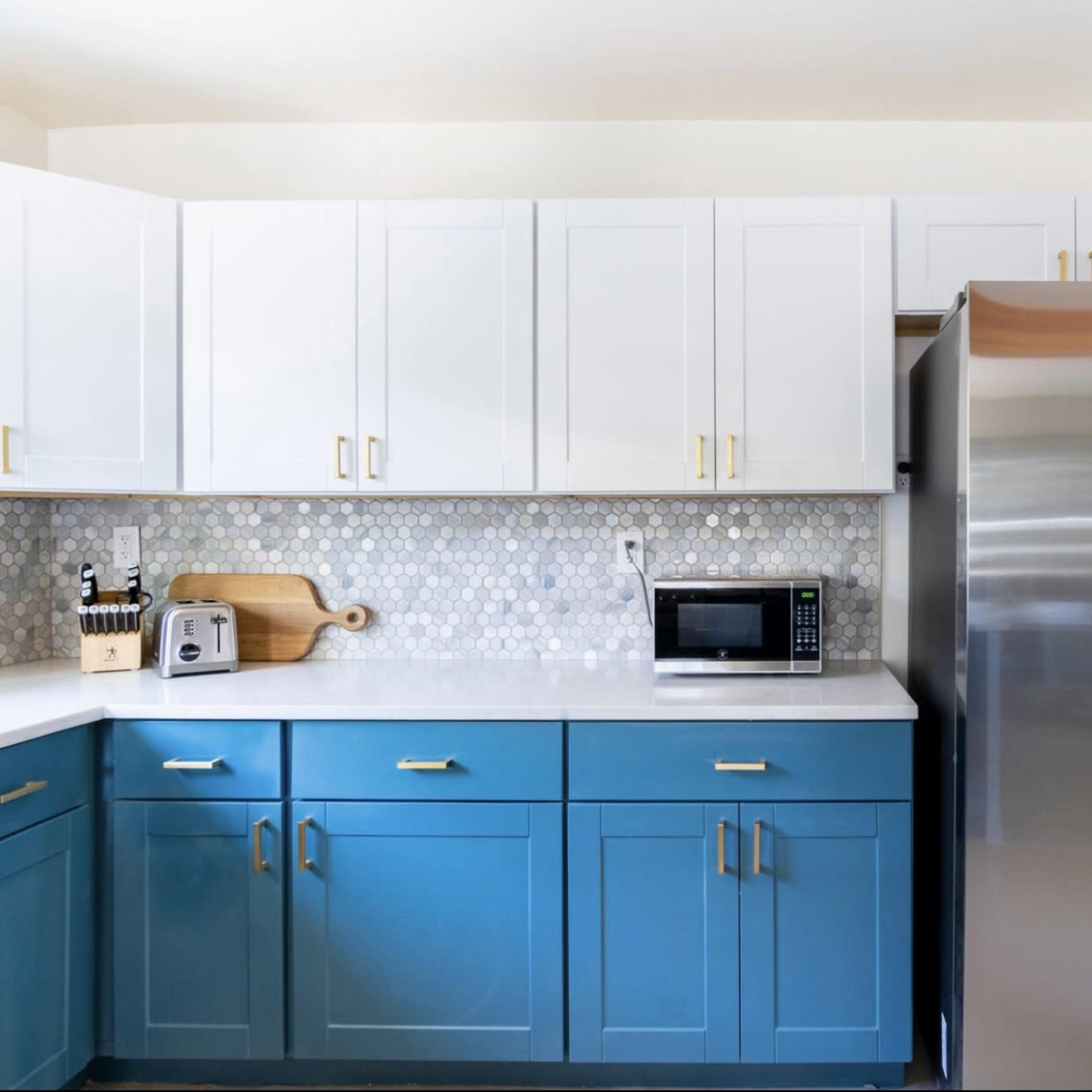 A modern kitchen with blue and white cabinets, a stainless steel refrigerator, a microwave, and a hexagon tile backsplash.