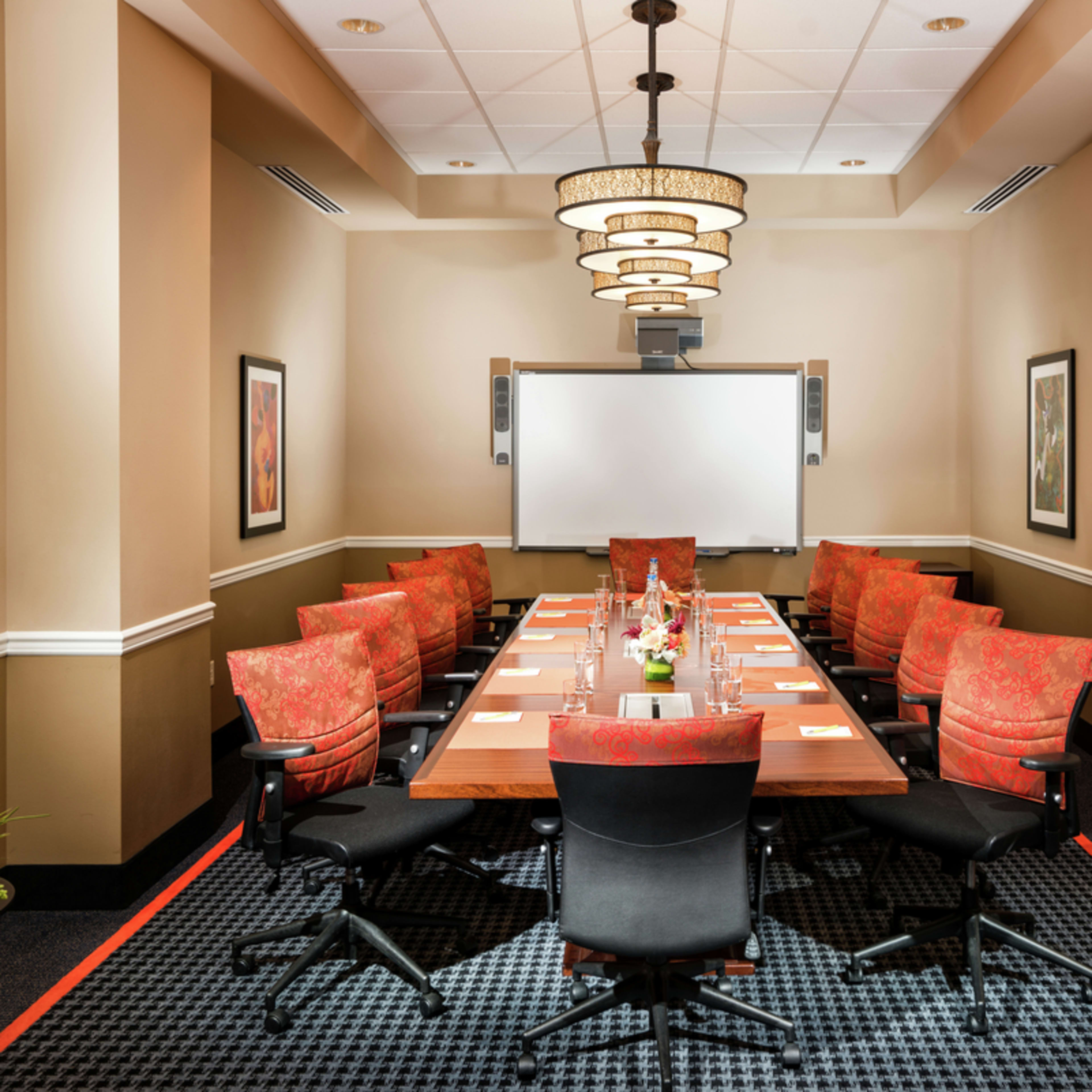 A conference room features a large rectangular table surrounded by upholstered chairs and a presentation screen at the front.