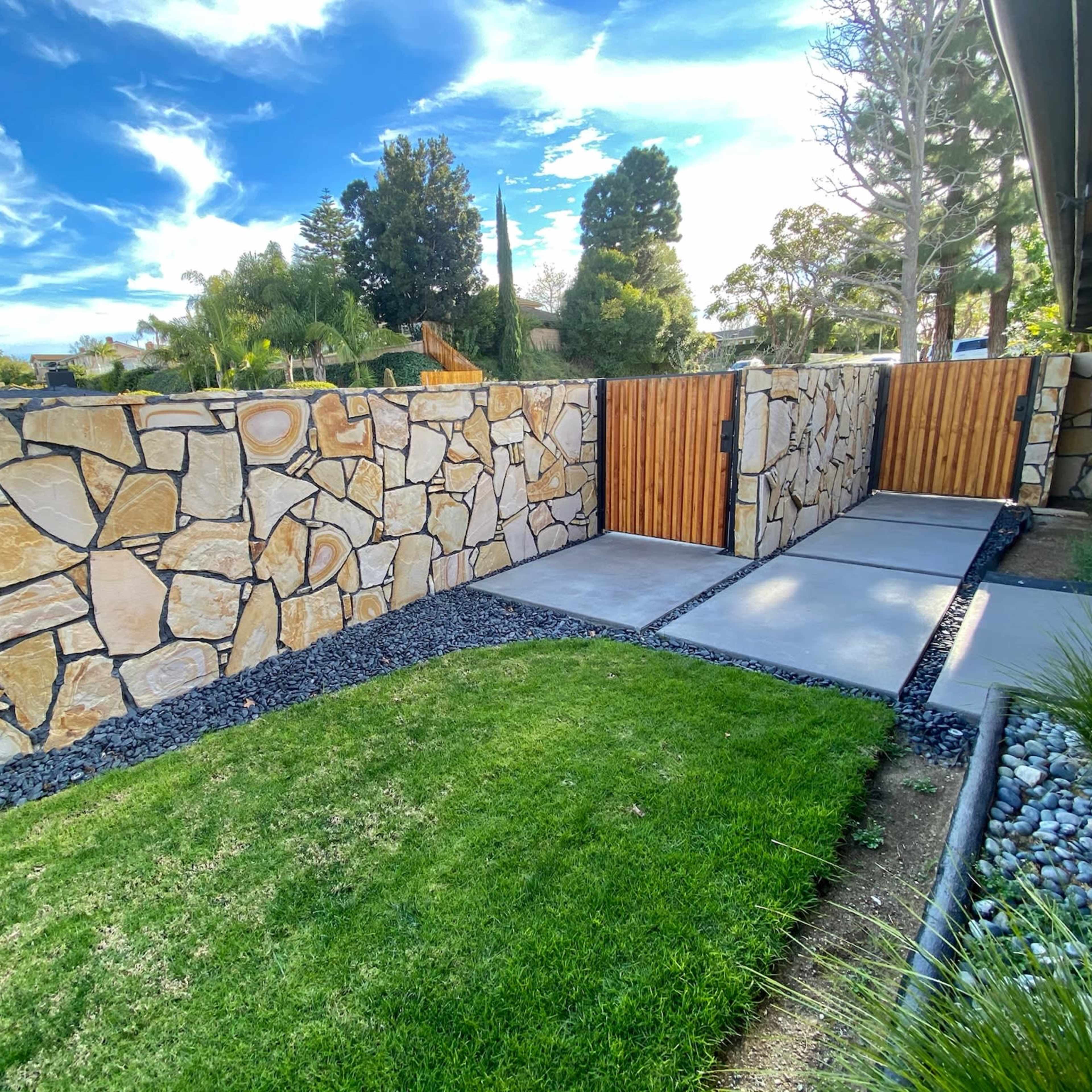 A landscaped yard features a stone wall with wooden gates and a concrete pathway bordered by grass and decorative gravel.