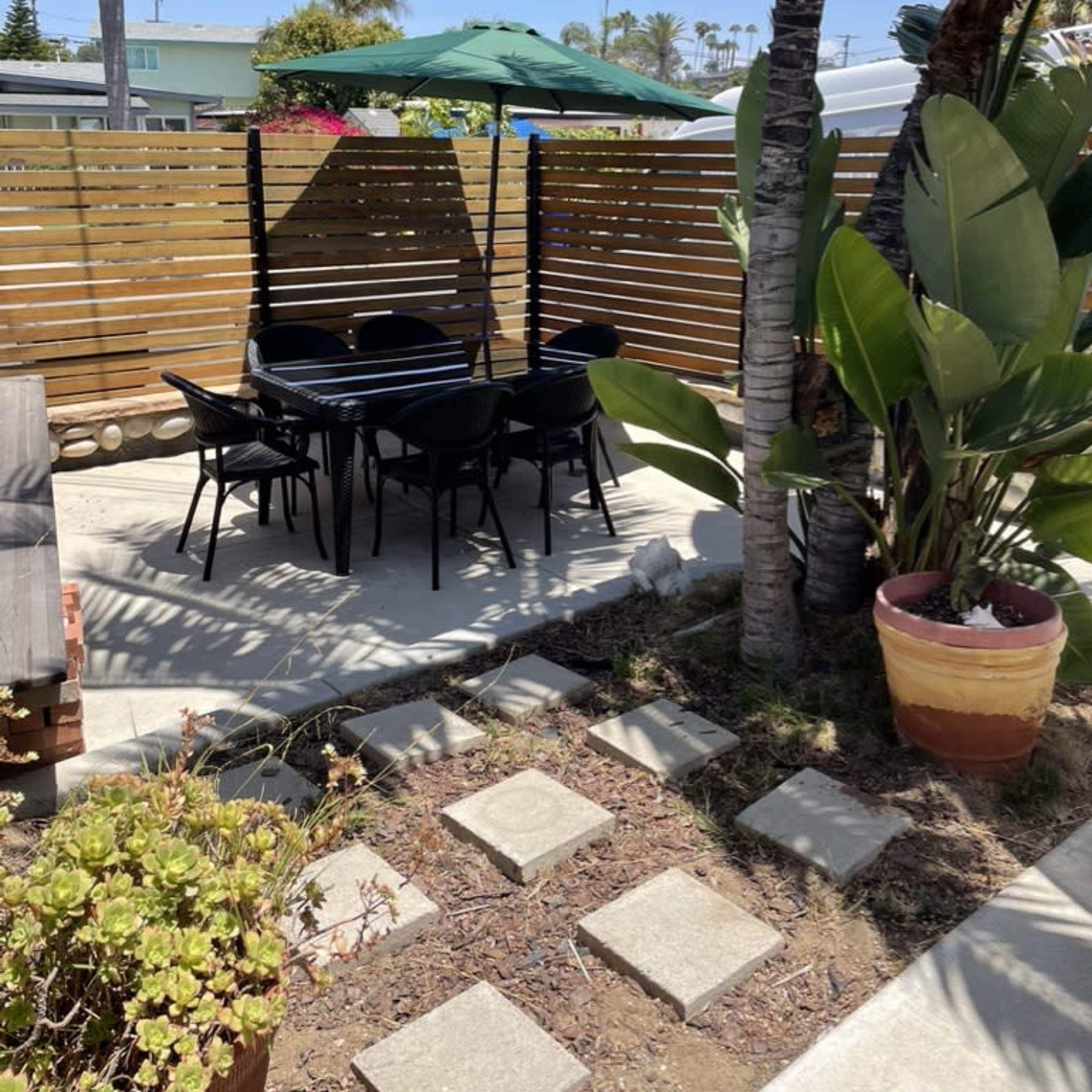 A patio area with a black table and chairs, shaded by a green umbrella, surrounded by wooden fencing and tropical plants.