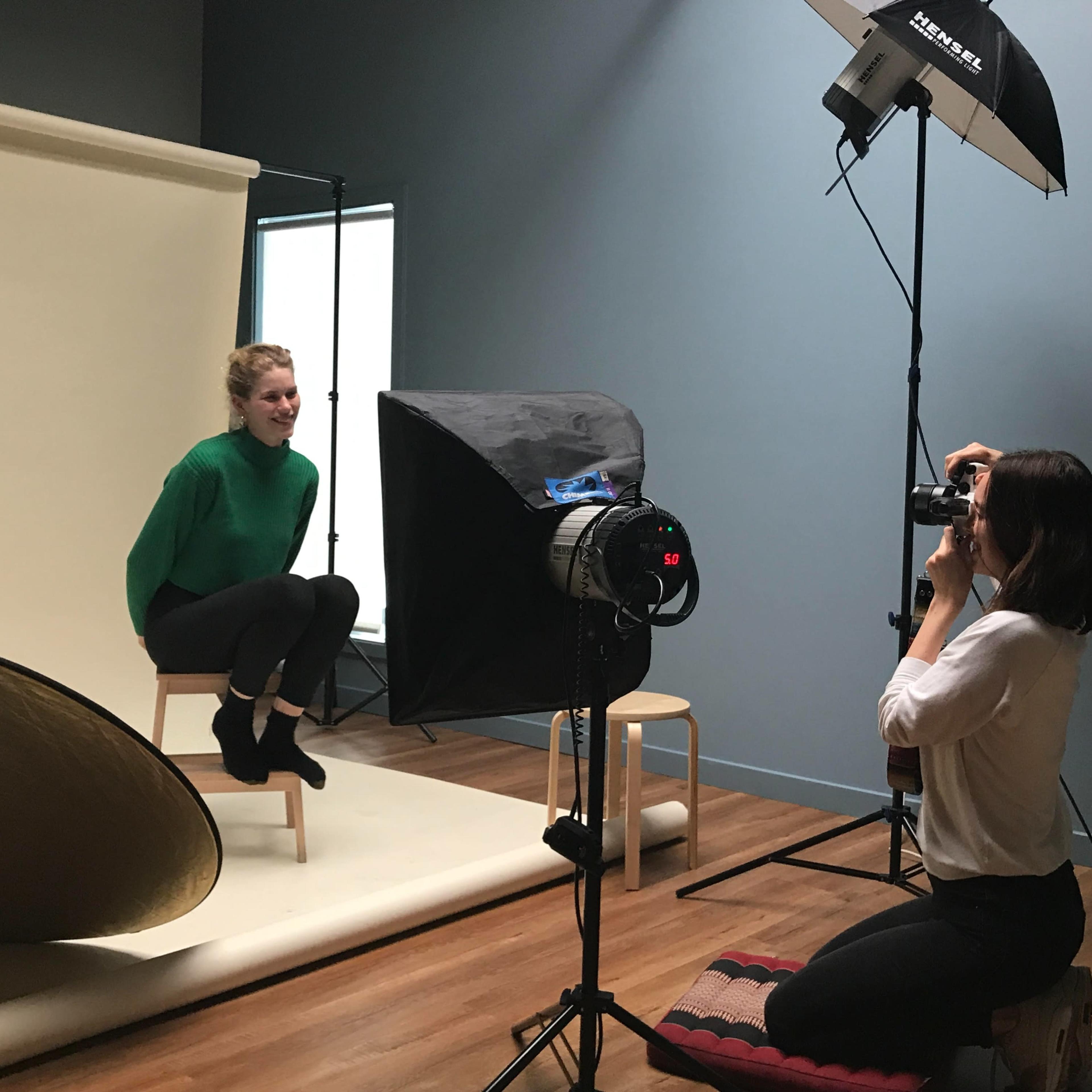 A photographer captures a portrait of a woman seated on a stool in a studio setting.