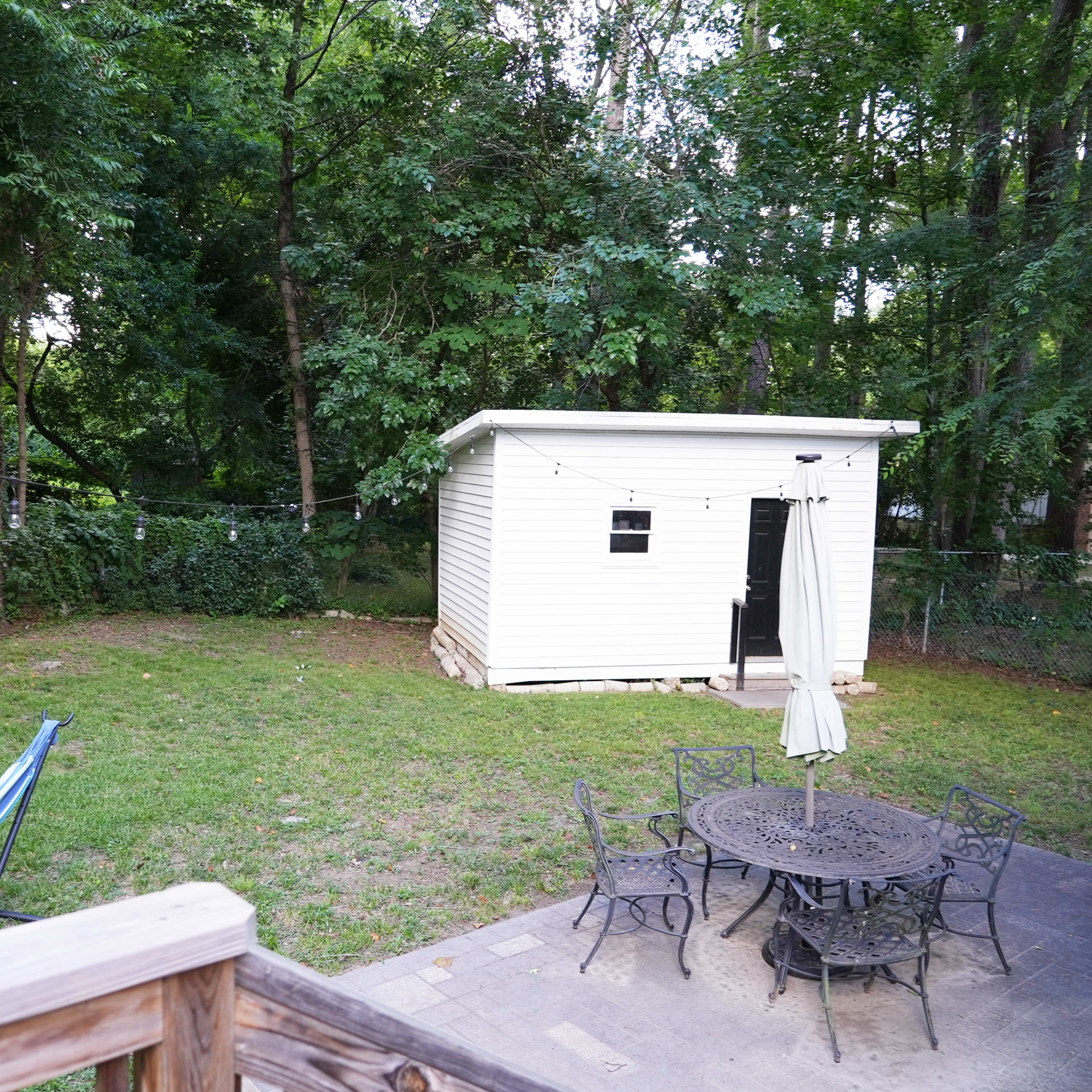A backyard with a white shed surrounded by trees, a hammock, and a circular table with chairs.