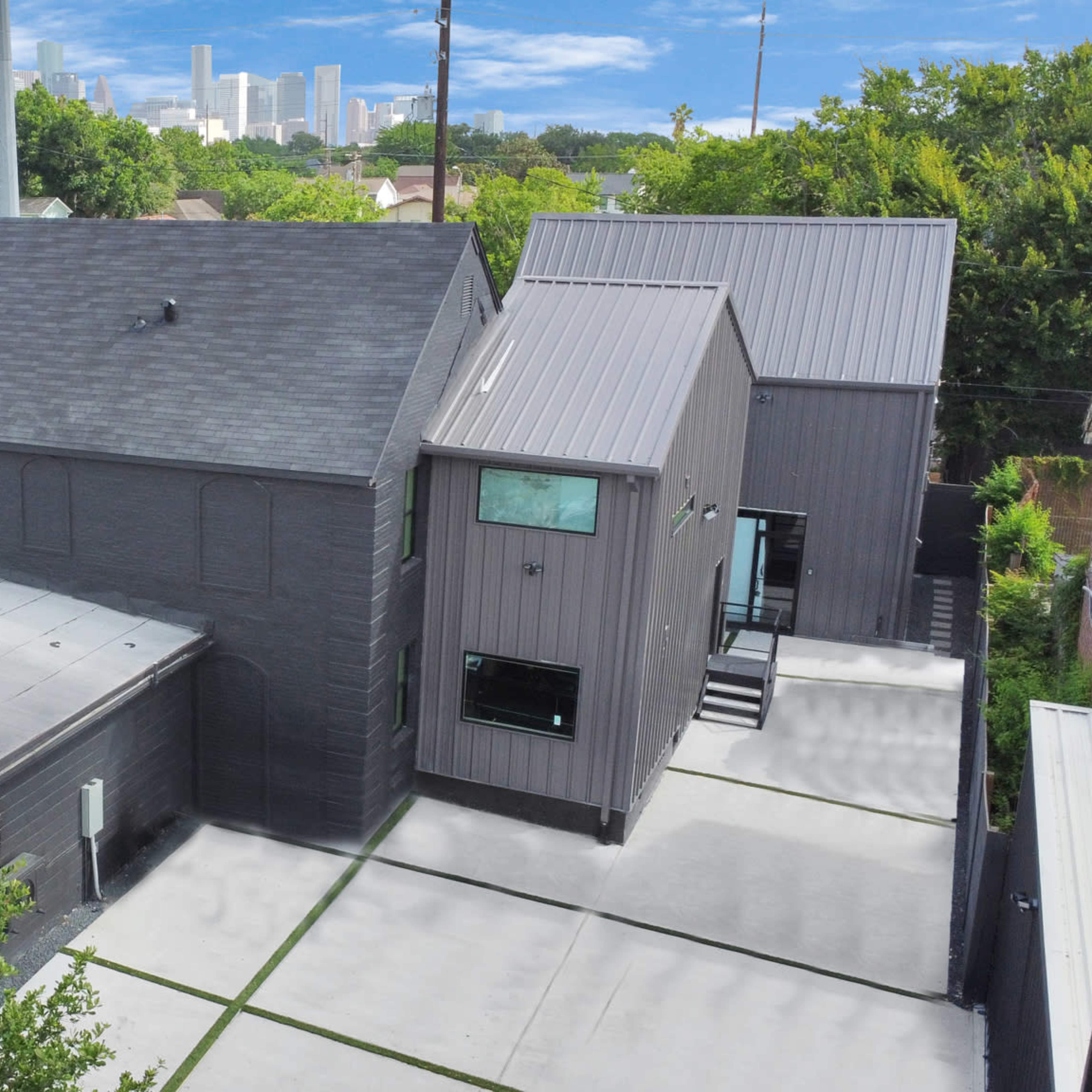 A modern residential building with a flat roof and angular design, surrounded by greenery and located near a city skyline in the background.