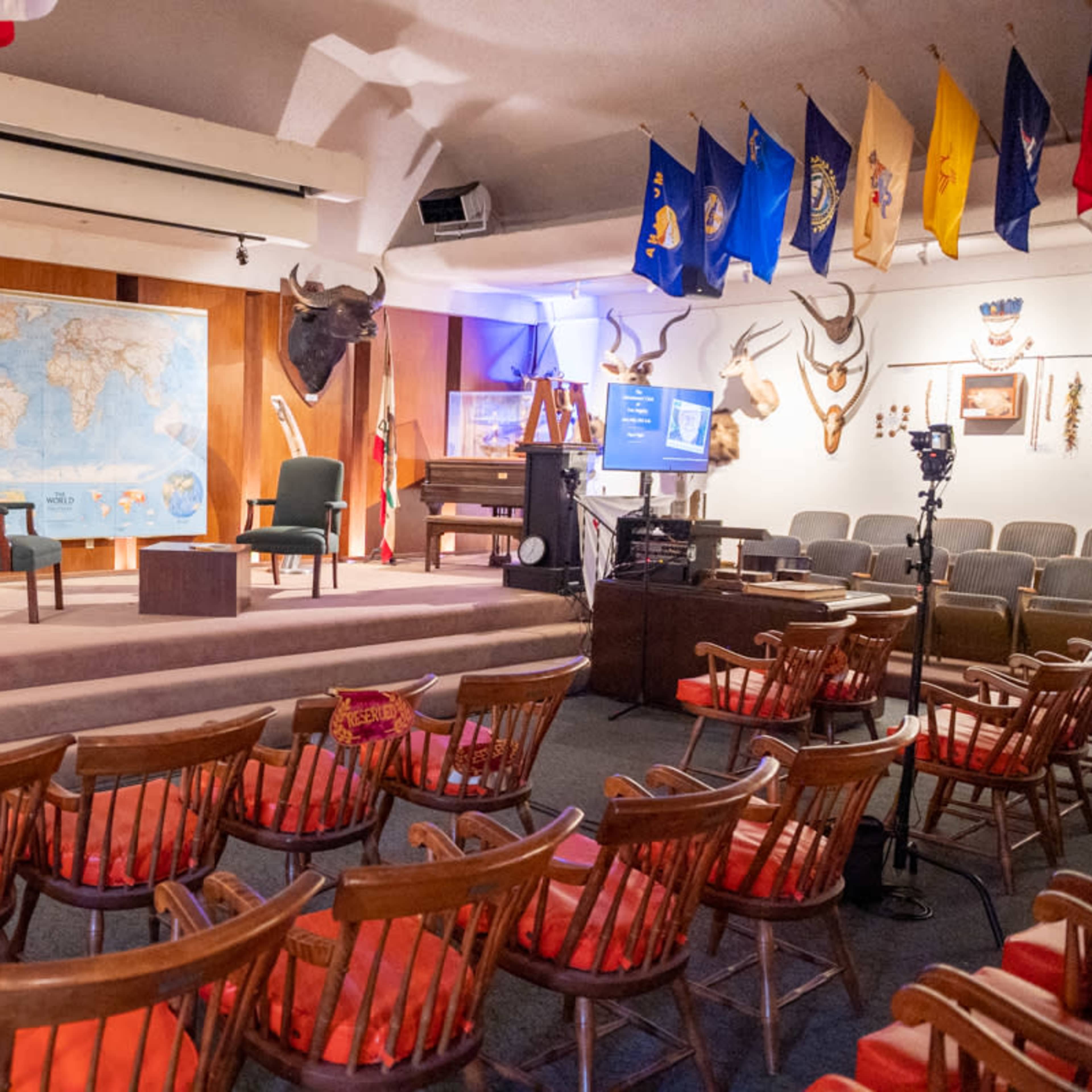 A stage setting with two chairs and a small table in front of a world map, surrounded by animal mounts and flags, with an arrangement of wooden chairs facing the stage.