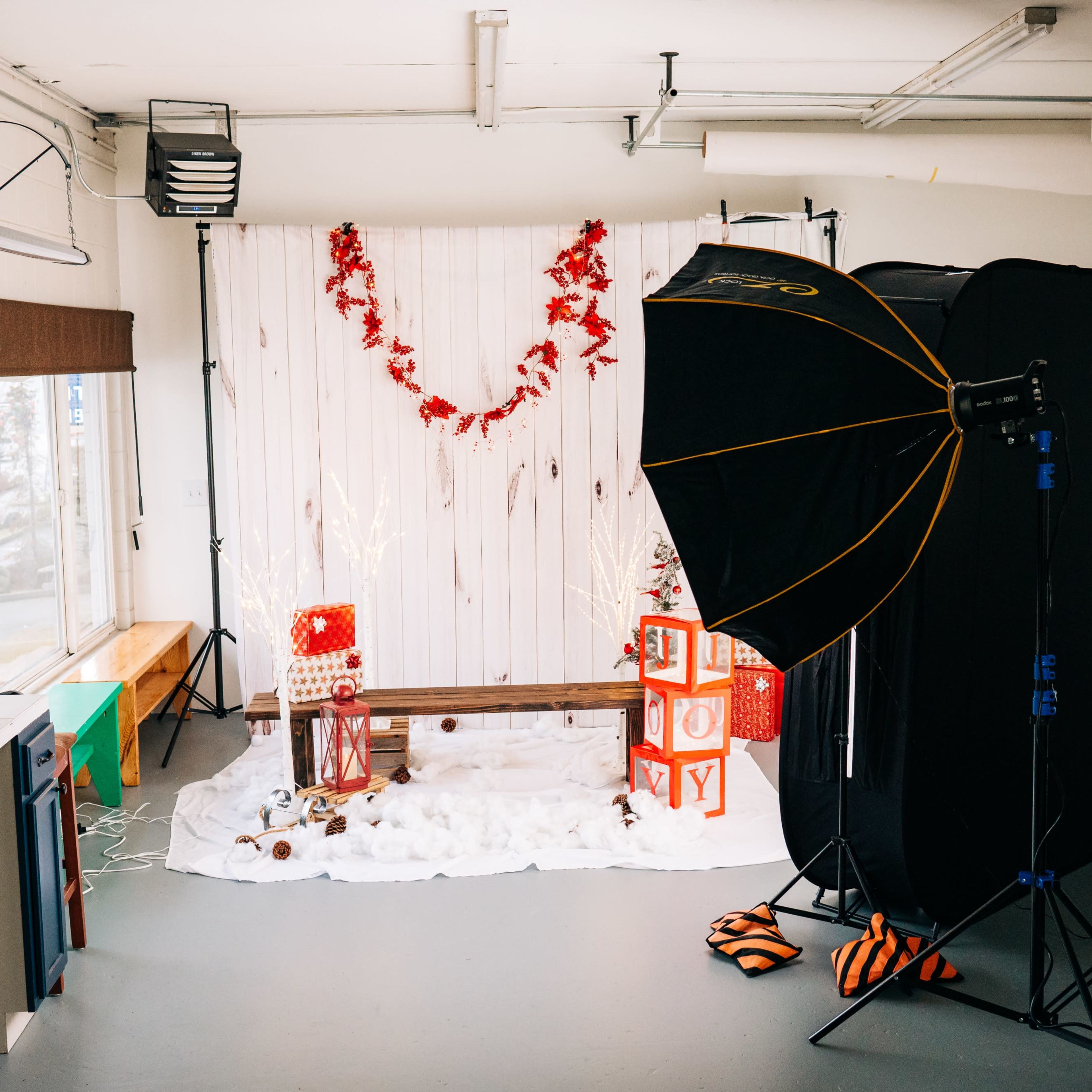 A photography studio is set up with a backdrop of white wooden panels adorned with red decorations, featuring a bench and Christmas-themed props.