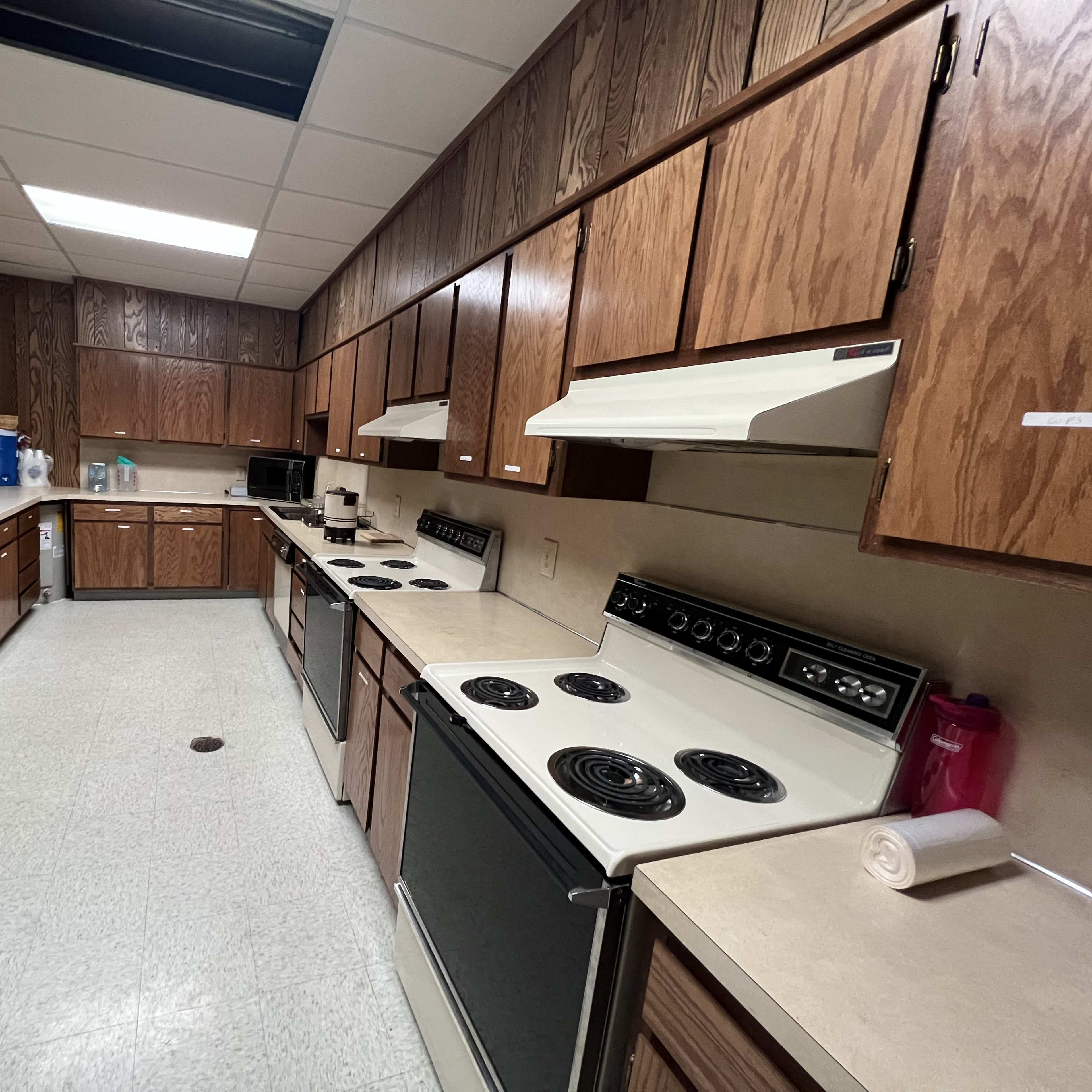 A long kitchen with wooden cabinets, multiple stoves, and countertops, featuring appliances along one wall and a door at the far end.