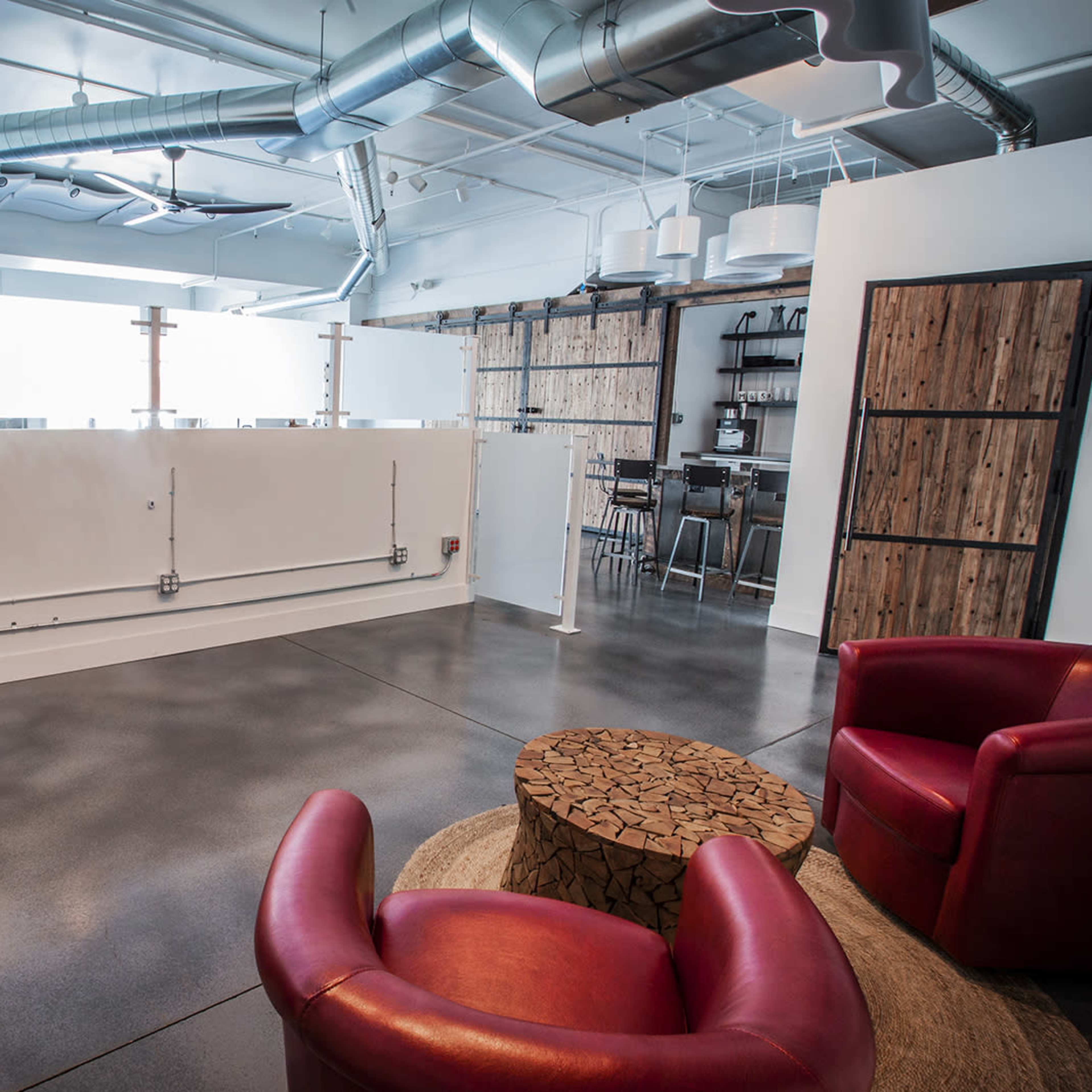 A modern office space with a seating area featuring two red chairs and a round wooden table, surrounded by industrial-style decor.