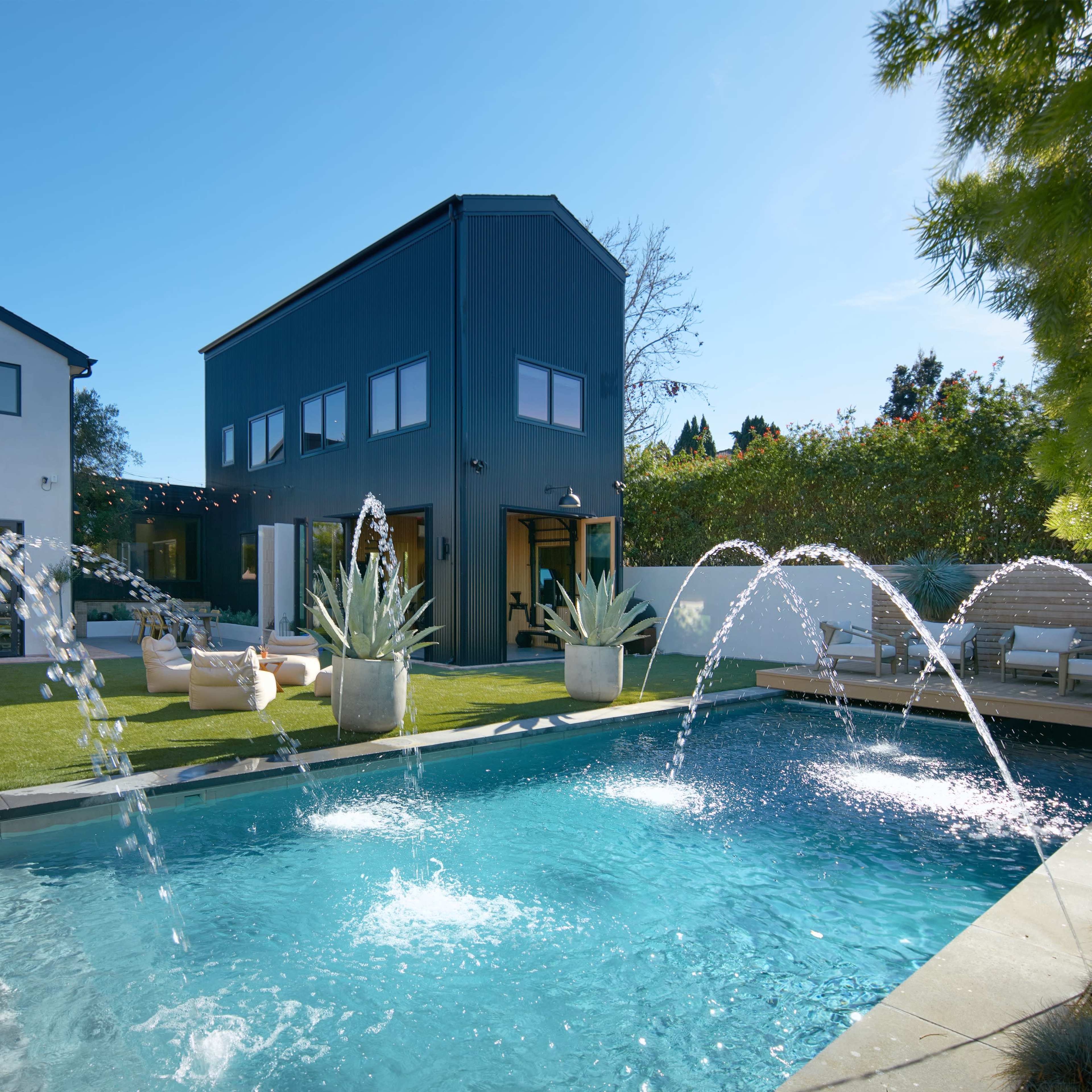 A modern backyard features a swimming pool with water fountains, surrounded by landscaped greenery and contemporary architecture.