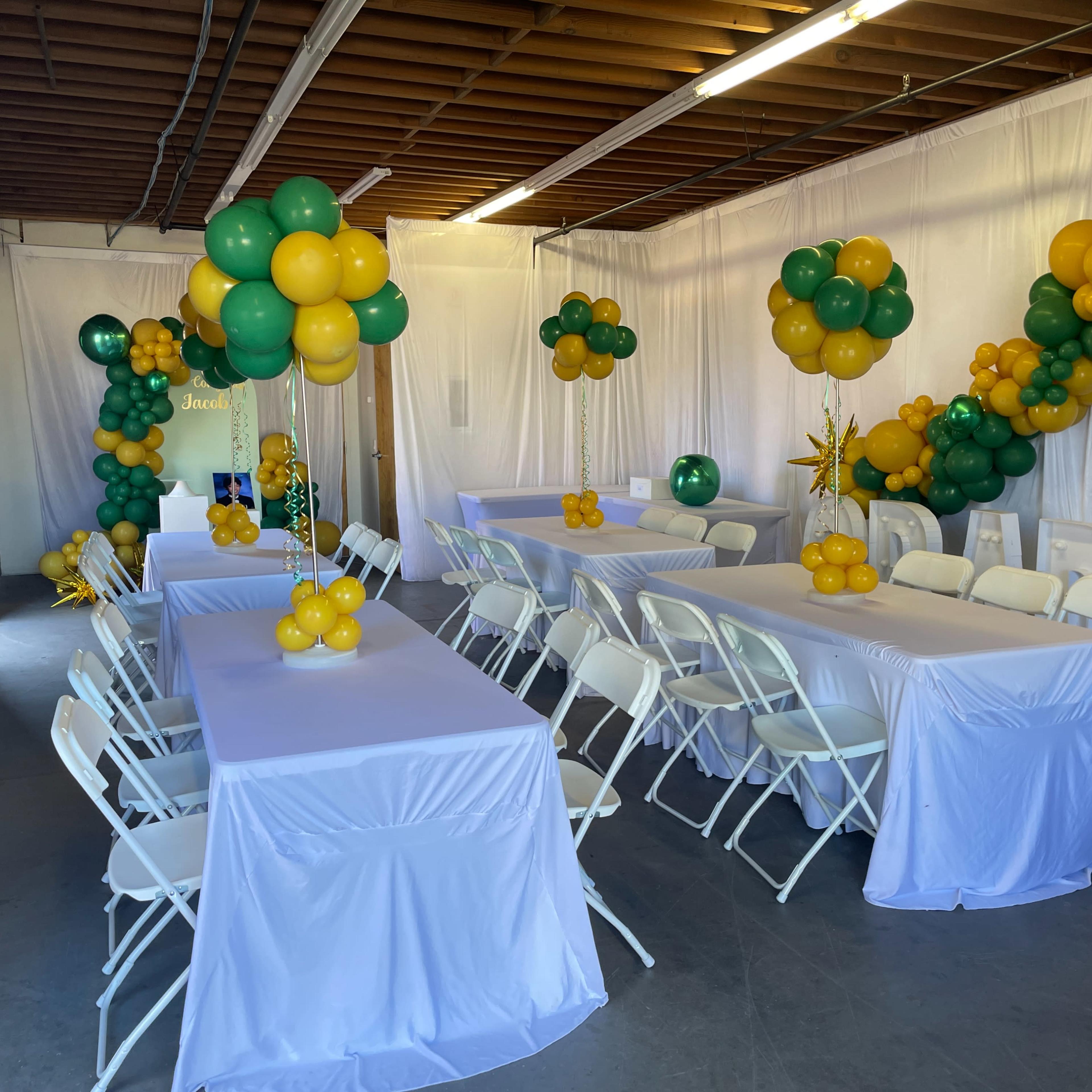 A decorated indoor space with rows of white tables covered with tablecloths, surrounded by white chairs, and embellished with green and yellow balloon arrangements.