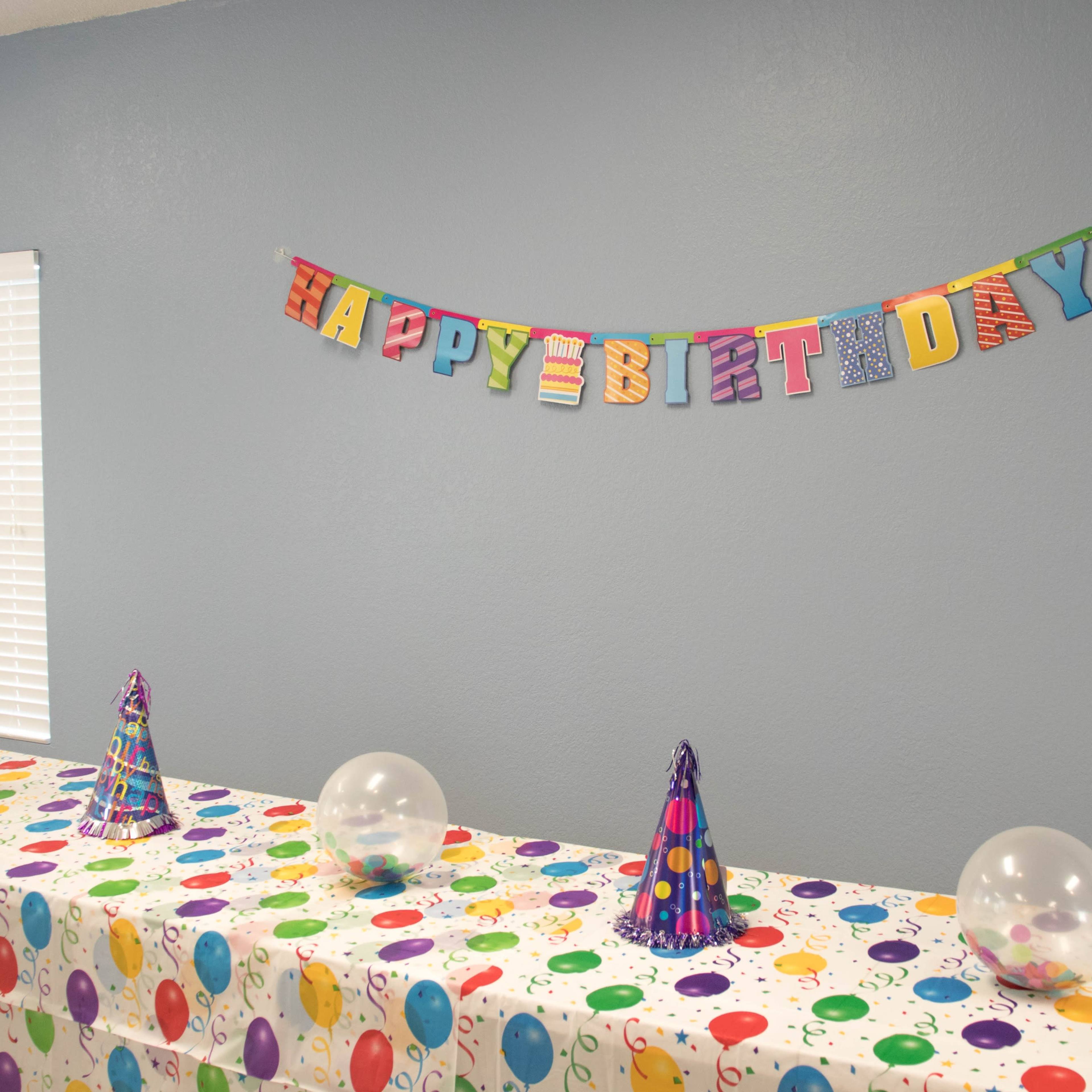 A decorated room features a "Happy Birthday" banner and a table adorned with a colorful, polka-dotted tablecloth and party hats.