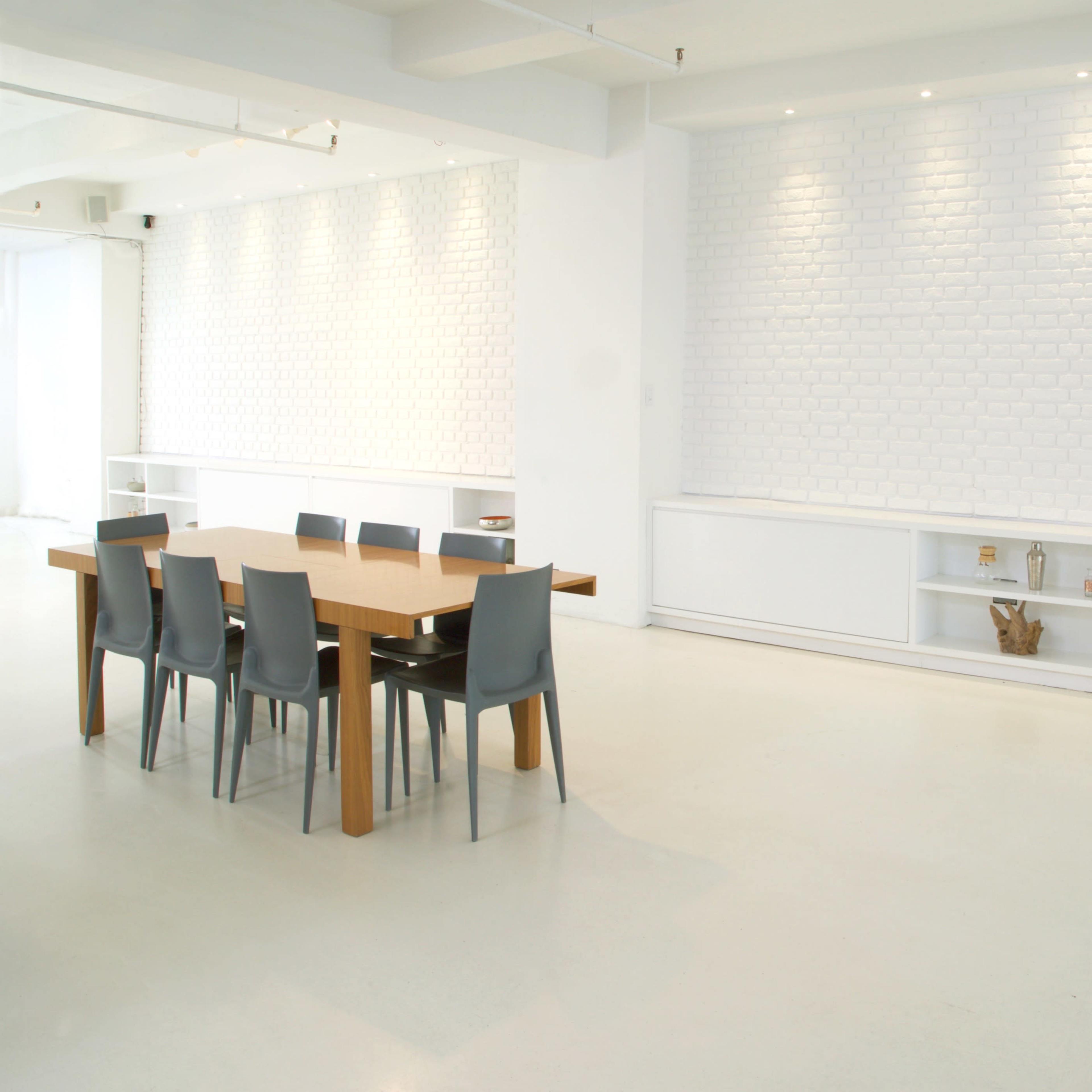A minimalist dining area features a wooden table surrounded by gray chairs, set against a white brick wall and a shelf displaying decorative items.