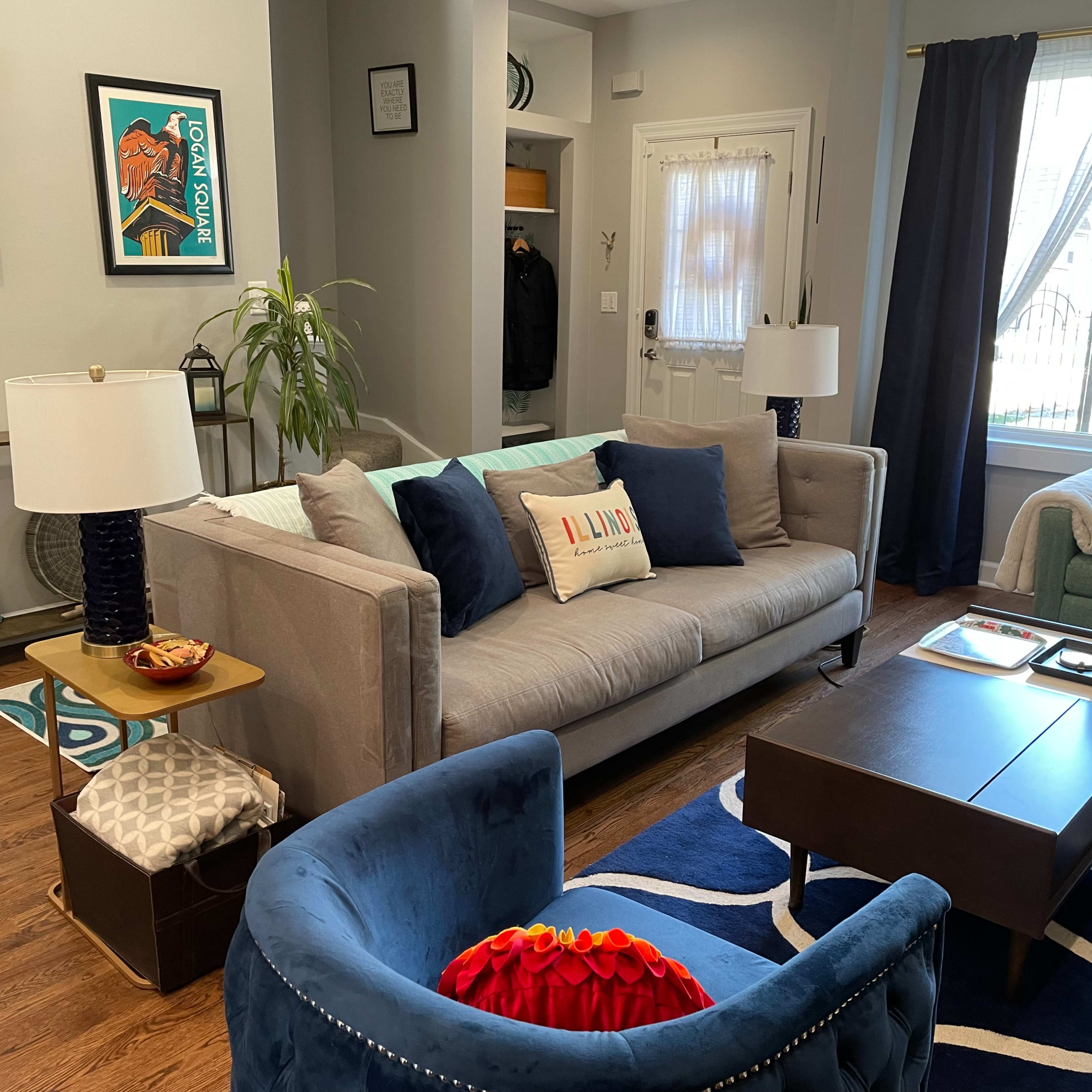 A cozy living room featuring a gray sofa with colorful cushions, a blue armchair, a wooden coffee table, and decorative artwork on the walls.