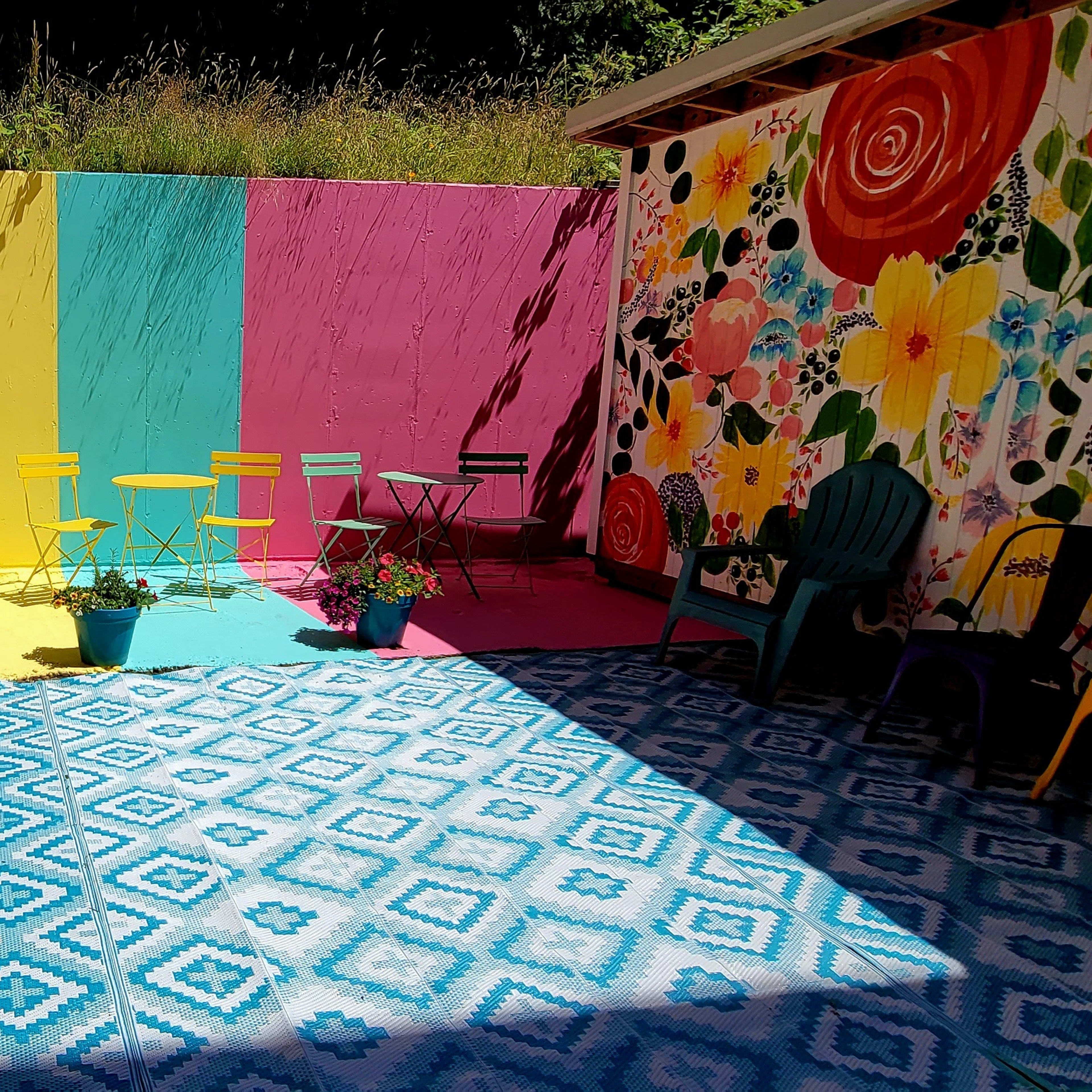 A vibrant outdoor space features colorful seating arranged along a striped mural, with a patterned rug and flowering plants enhancing the scene.