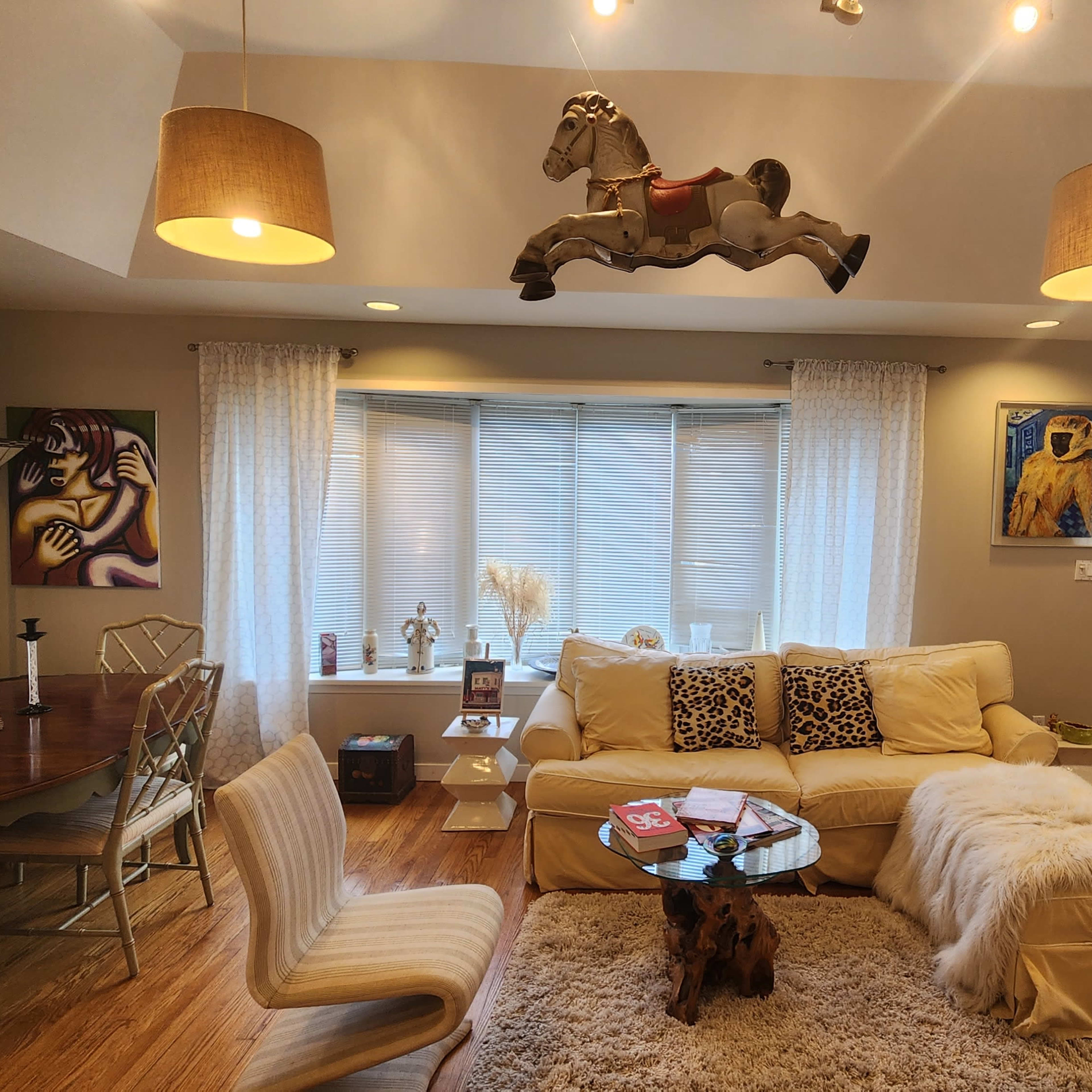 A spacious living room features a cream-colored sectional sofa, a wooden dining table, and a mounted horse sculpture above a window displaying sheer curtains.