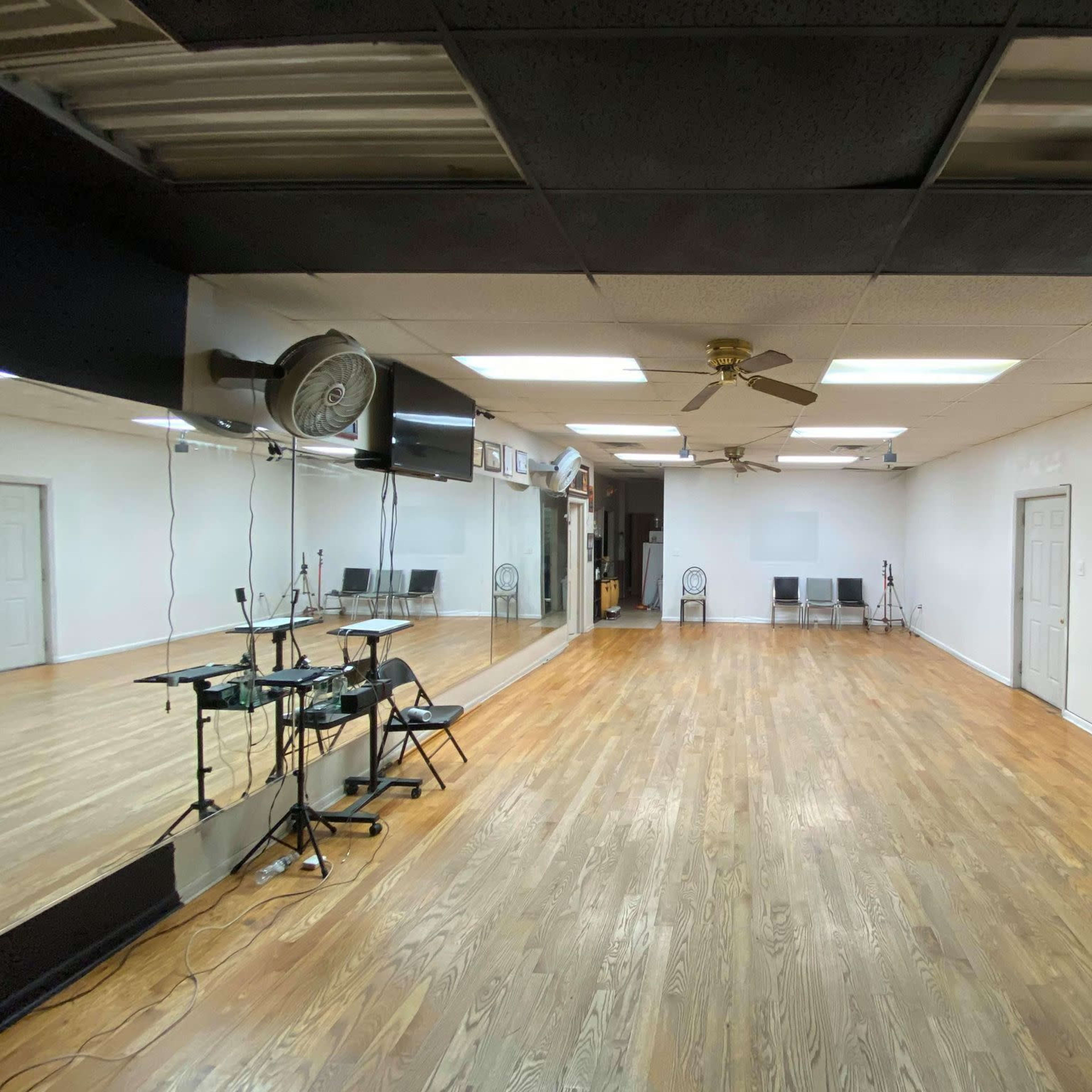 An empty room with wooden flooring, a ceiling fan, mirrors on one wall, and several chairs against the opposite wall.