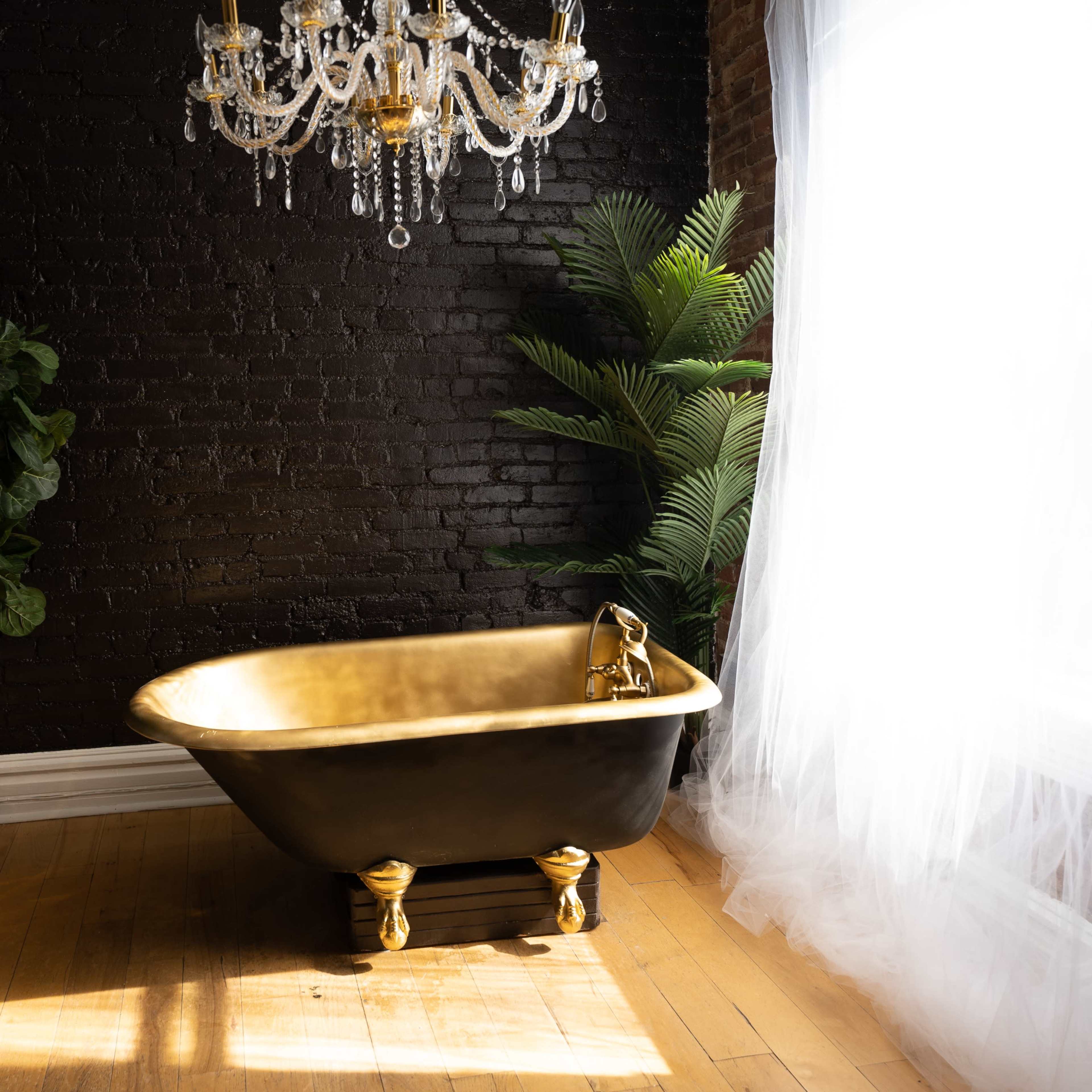 A black and gold freestanding bathtub is situated on hardwood flooring near a large window, surrounded by plants and a crystal chandelier.