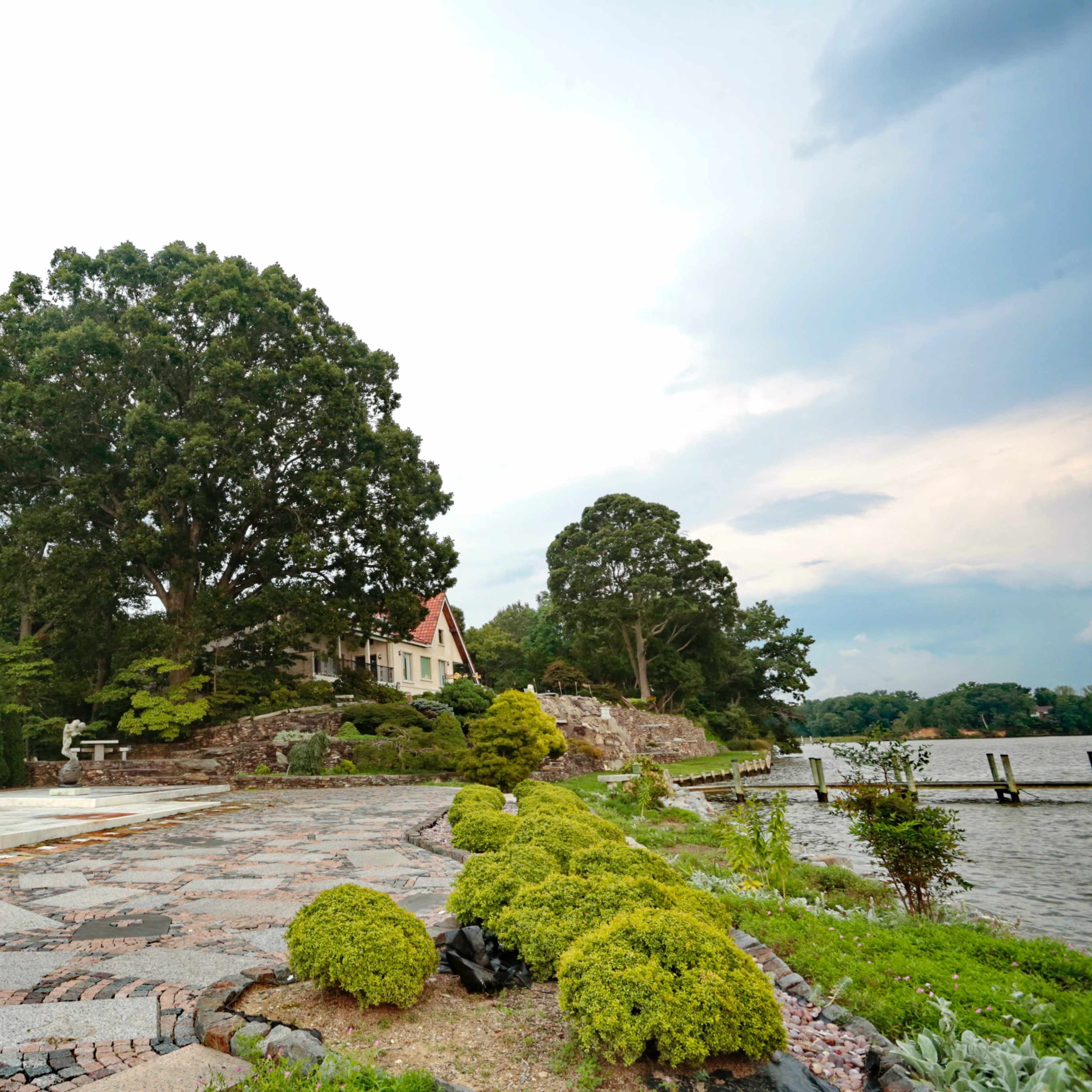 A landscaped waterfront area featuring a stone path, trimmed bushes, a statue, and a house set against a tree-lined background.