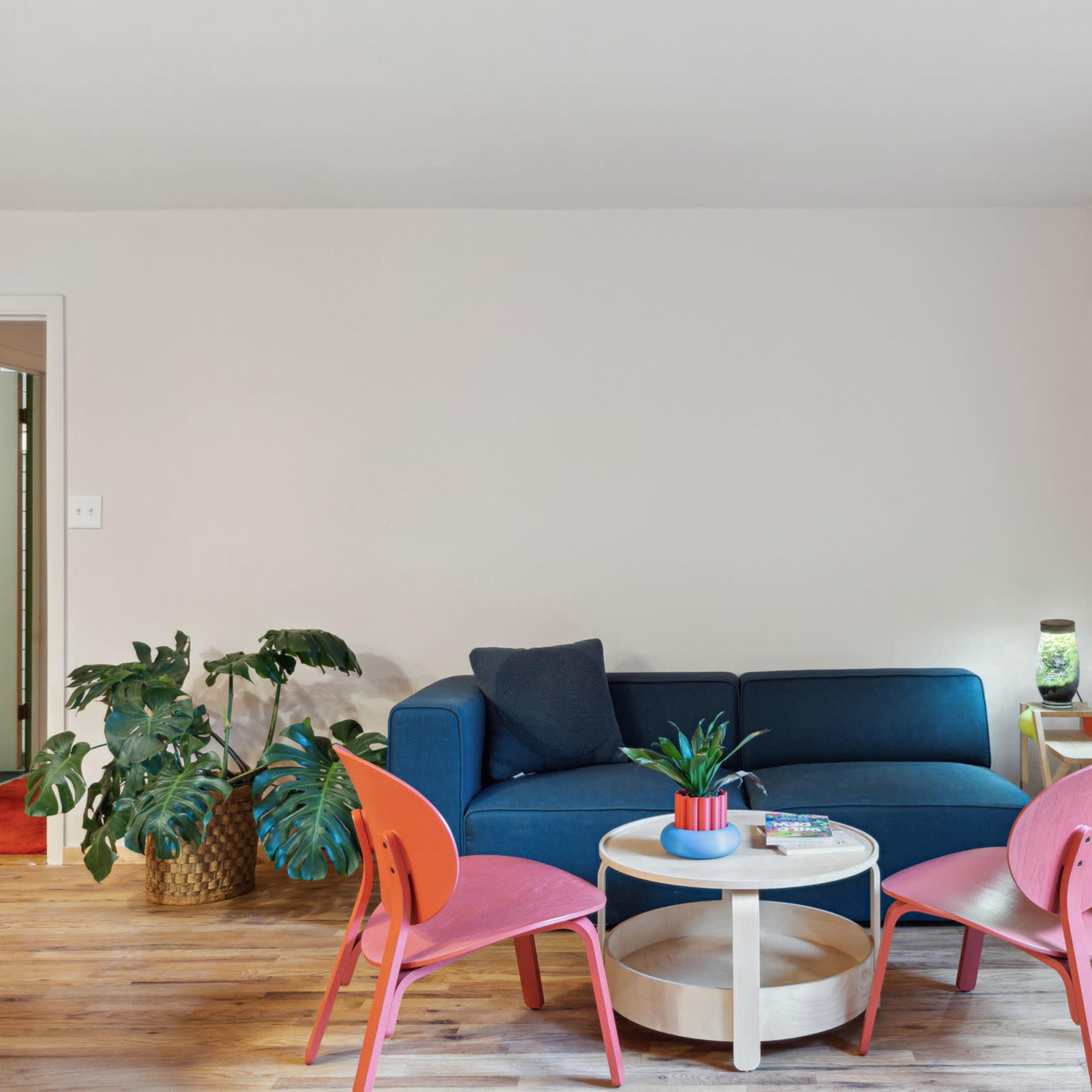 A blue couch is positioned against a light-colored wall, accompanied by a round coffee table and three pink chairs, with a potted plant and a doorway visible in the background.