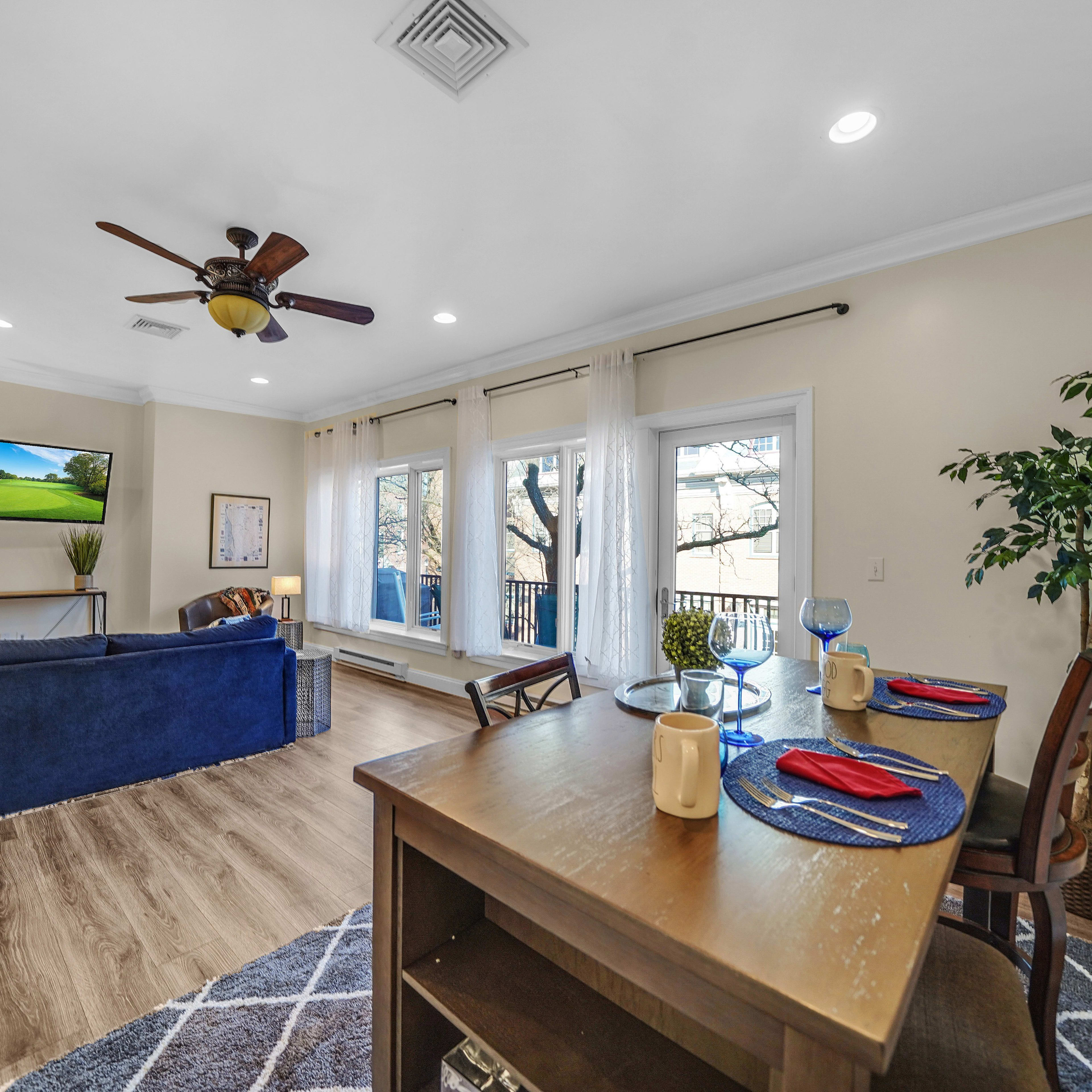 A well-lit living room with a blue sofa, a dining table set for two, and large windows overlooking a view of trees.