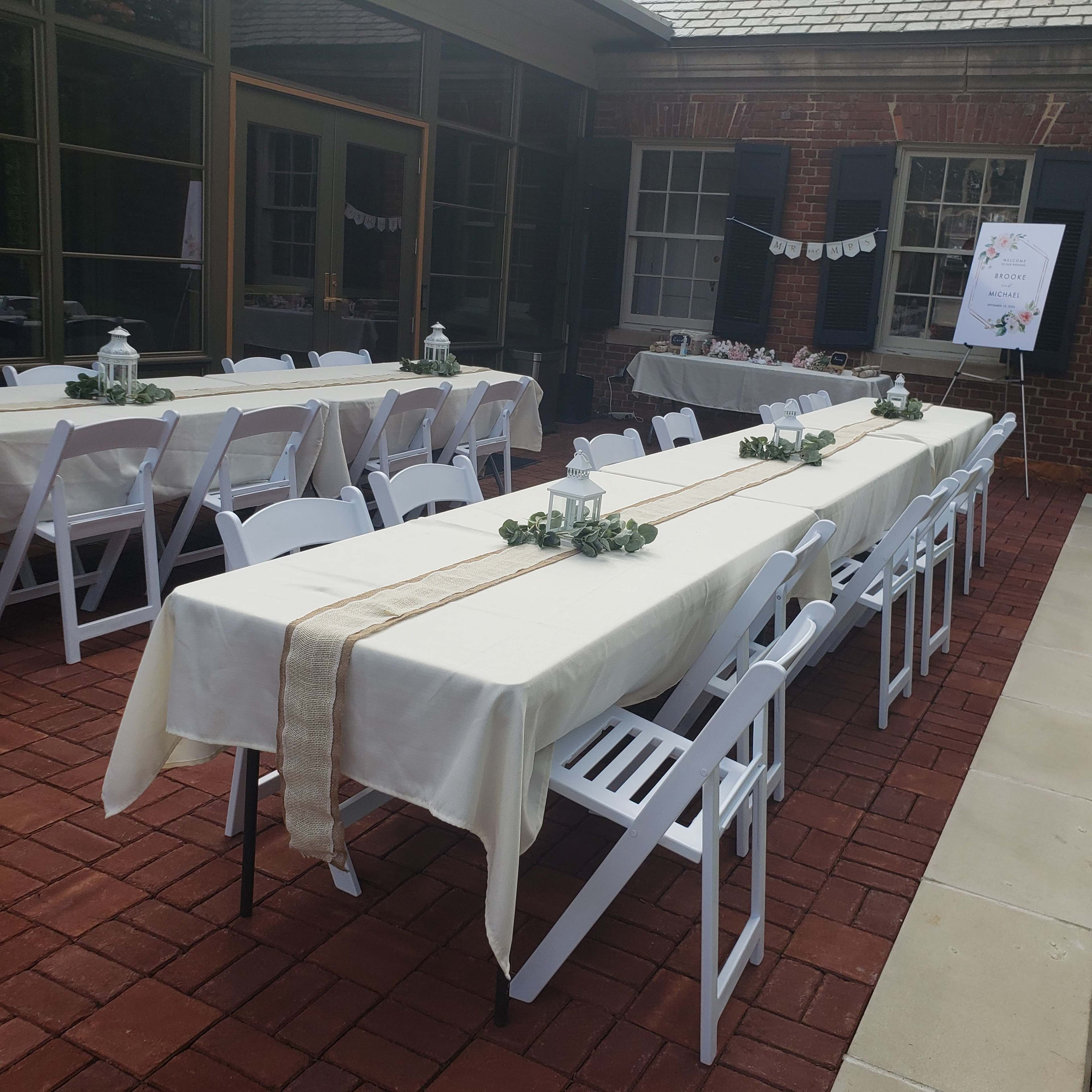A series of long tables with white tablecloths and decorative centerpieces are set up outdoors on a brick patio.