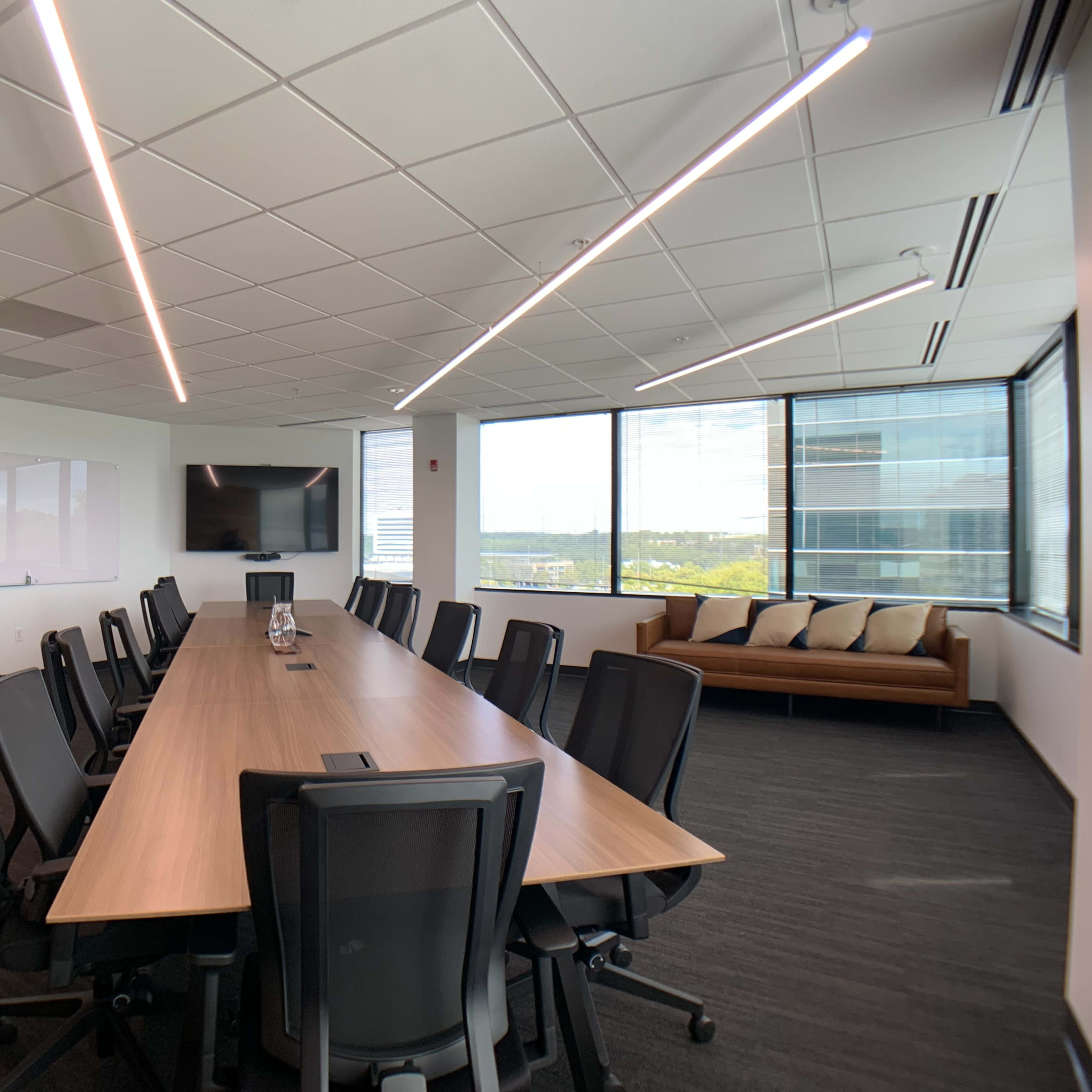 A modern conference room features a long wooden table surrounded by black chairs, with large windows offering a view of the outside.
