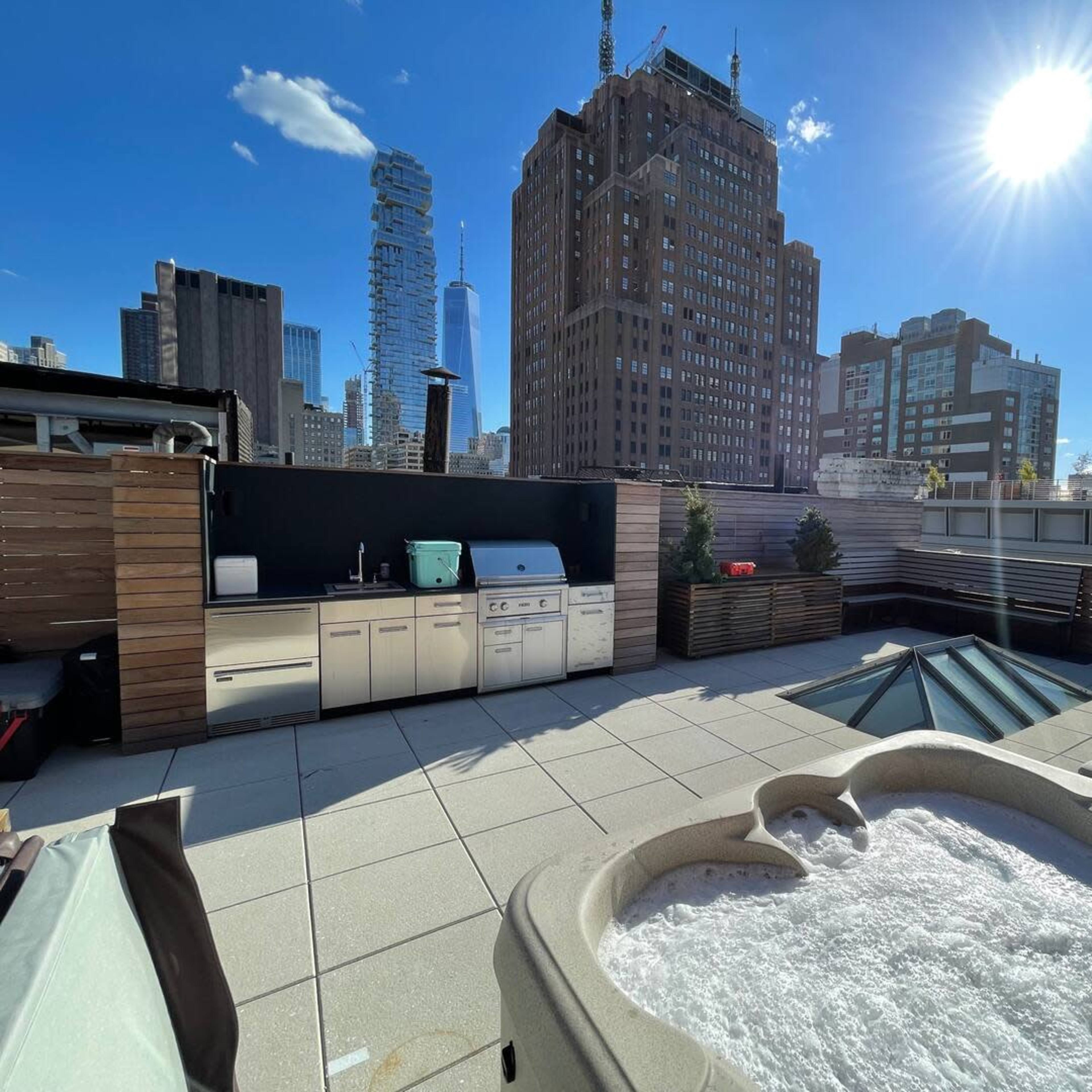 A rooftop space featuring a hot tub, an outdoor kitchen area, and a view of tall city buildings under bright sunlight.