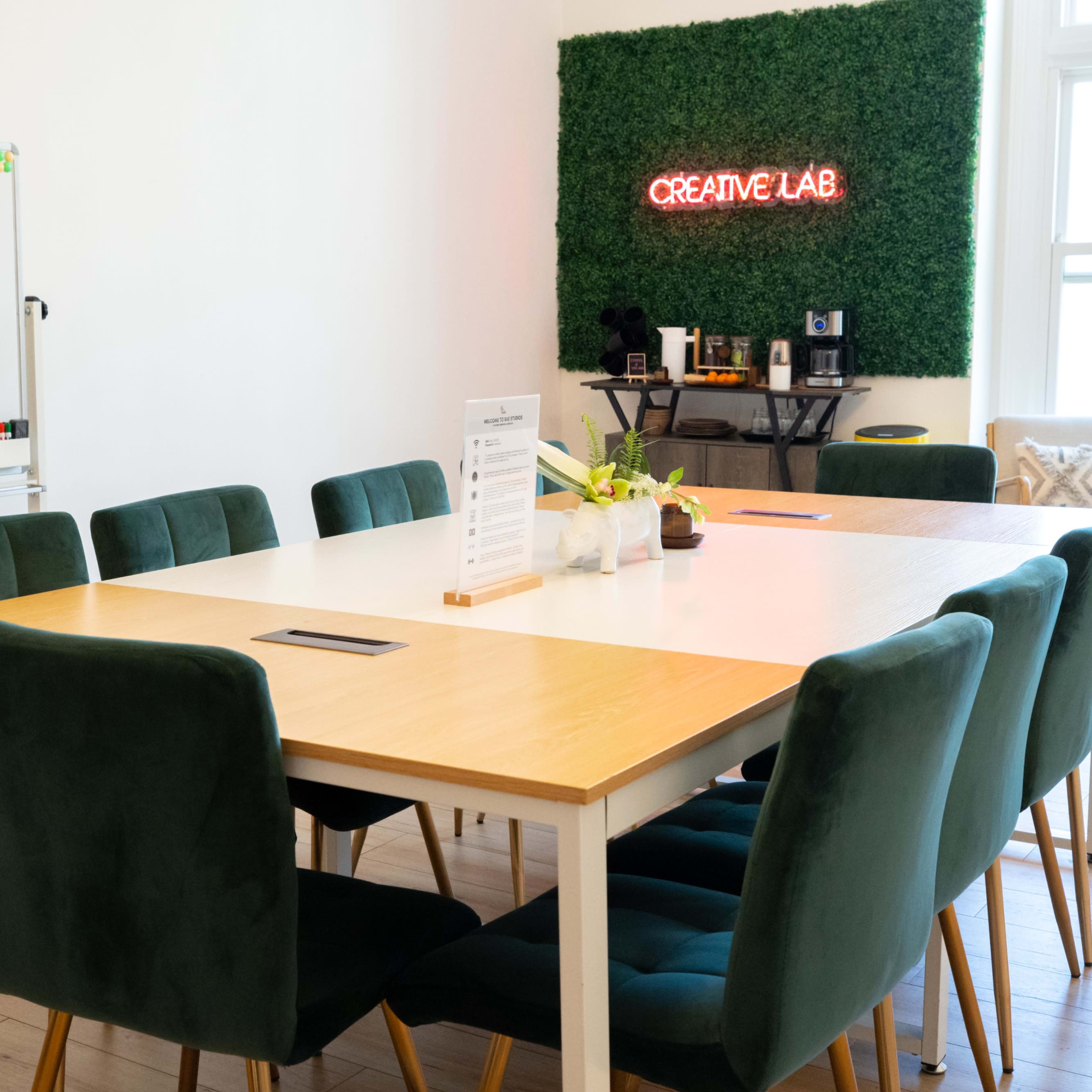 A large wooden table with green chairs is arranged in a bright room featuring a "CREATIVE LAB" sign made of greenery on the wall.