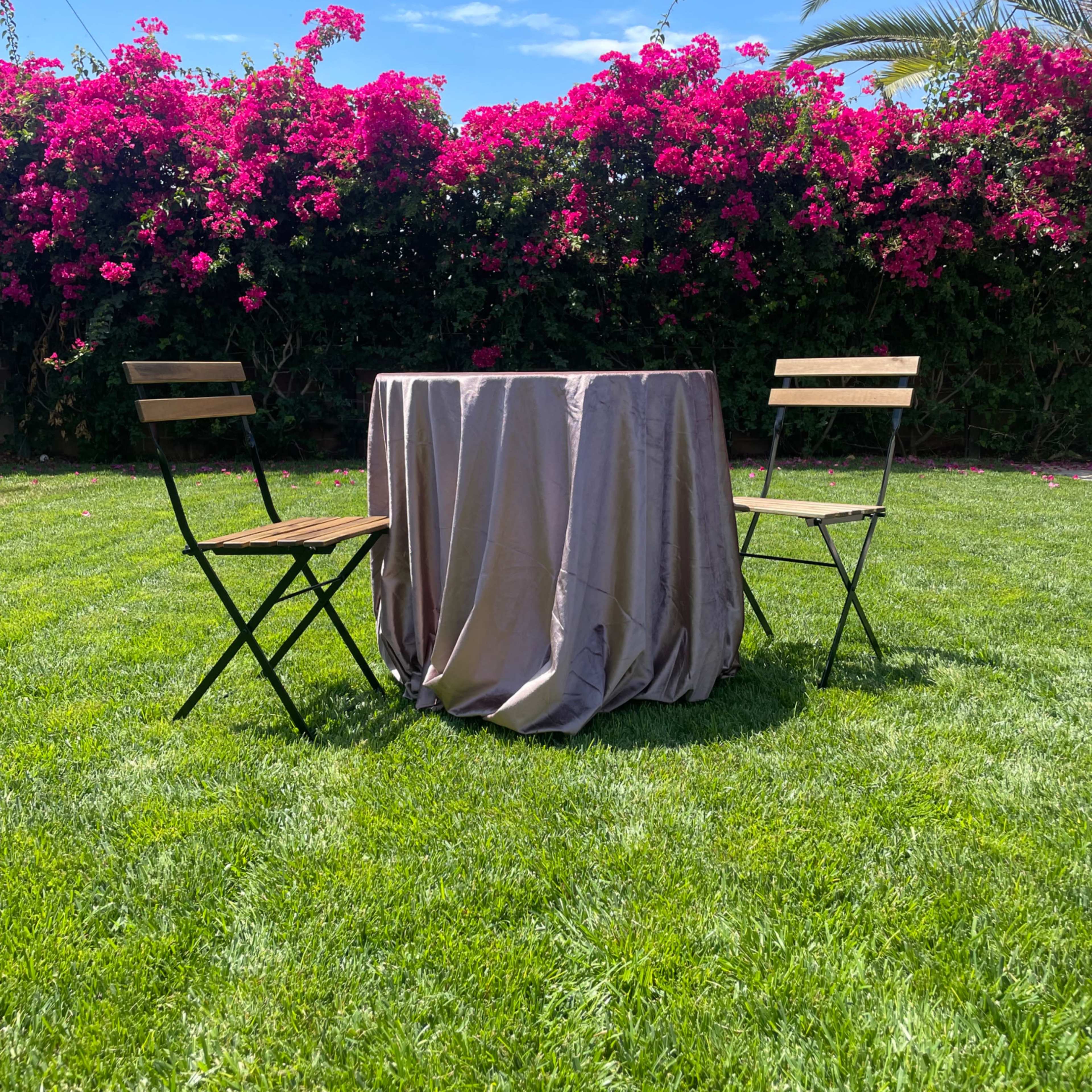 A round table with a tablecloth is set between two chairs on a grassy area, surrounded by blooming pink flowers and palm trees.