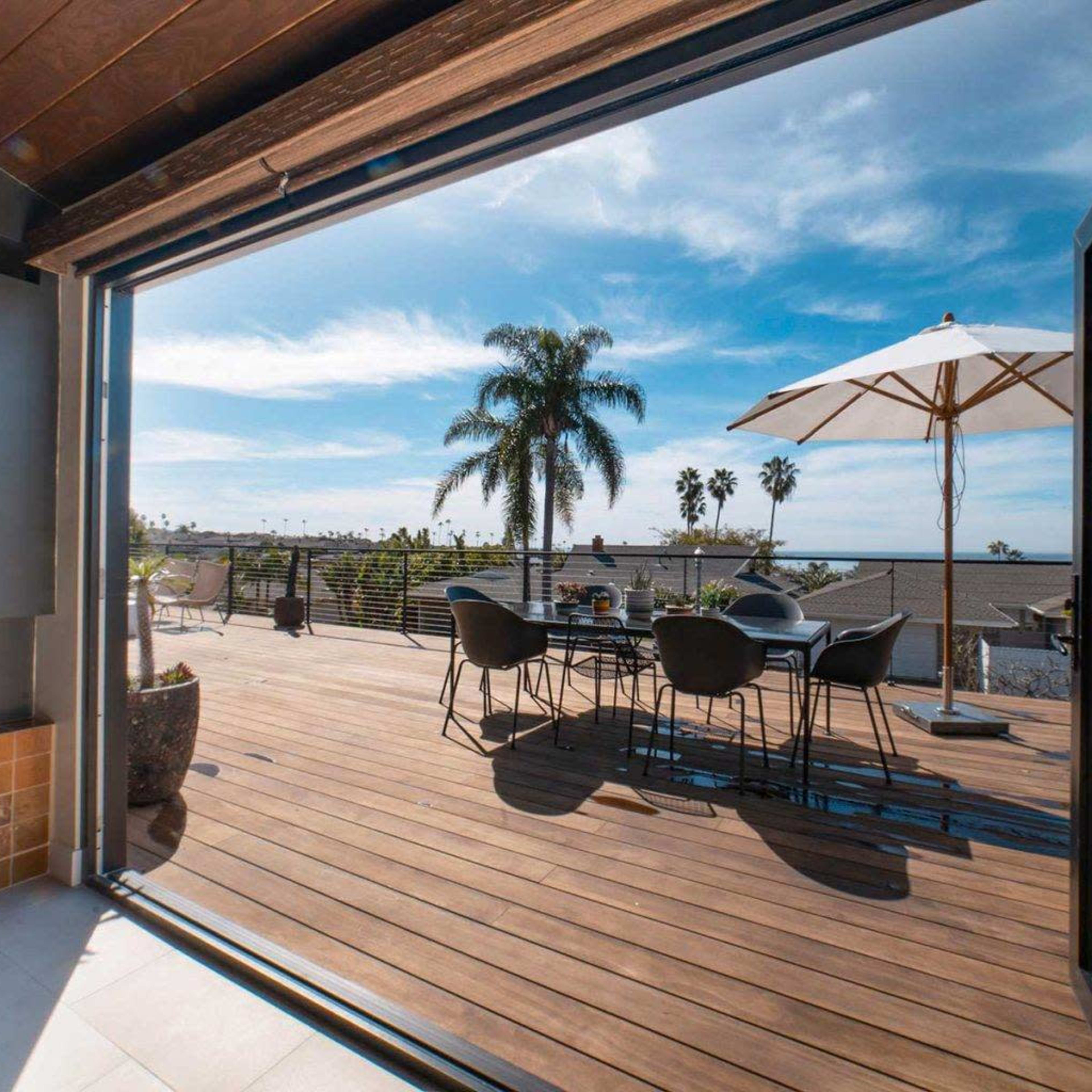 A spacious wooden deck with a dining table and chairs, surrounded by palm trees and an umbrella, under a bright blue sky.