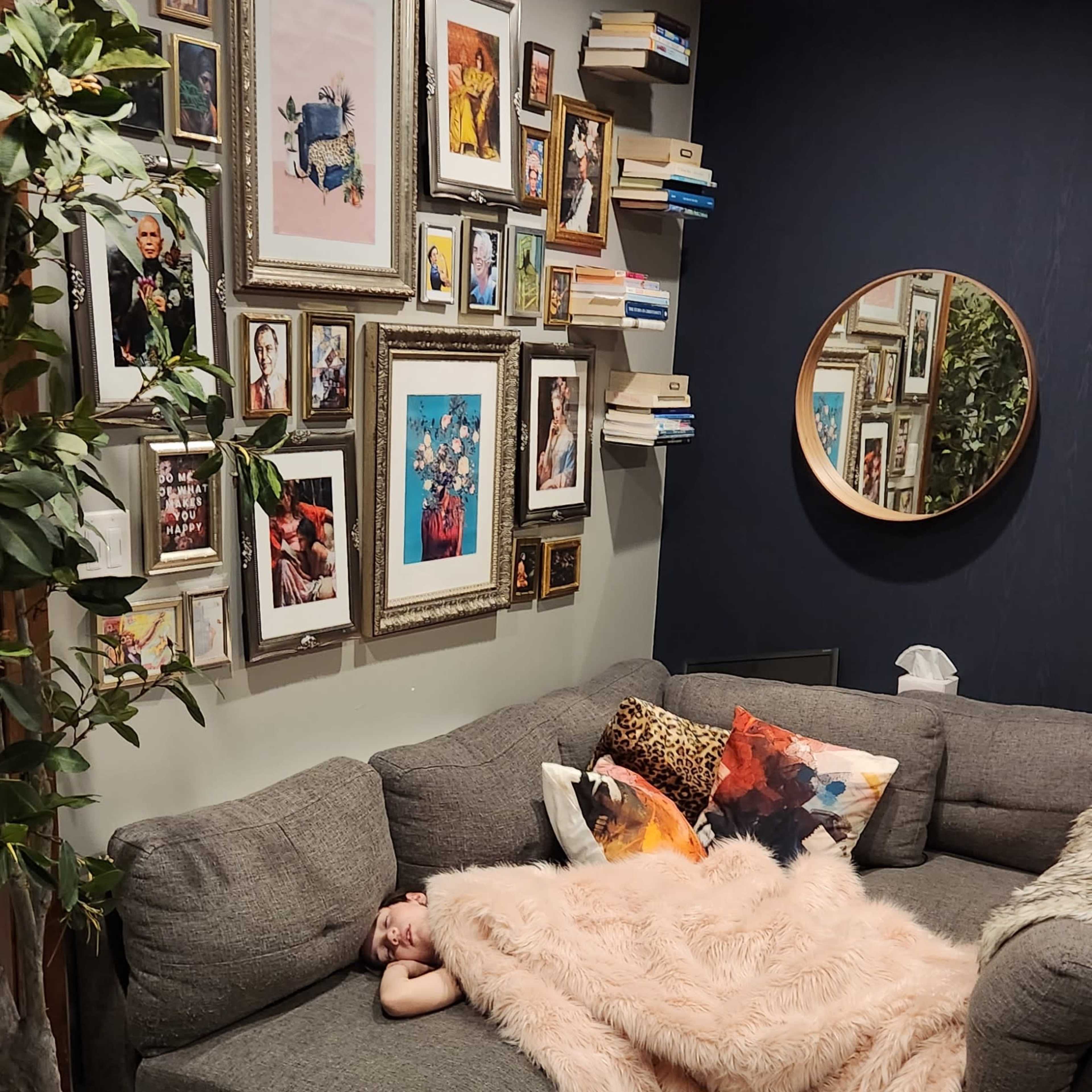 A person is sleeping on a gray sectional sofa draped with a pink blanket, surrounded by a gallery wall of framed photos and books.