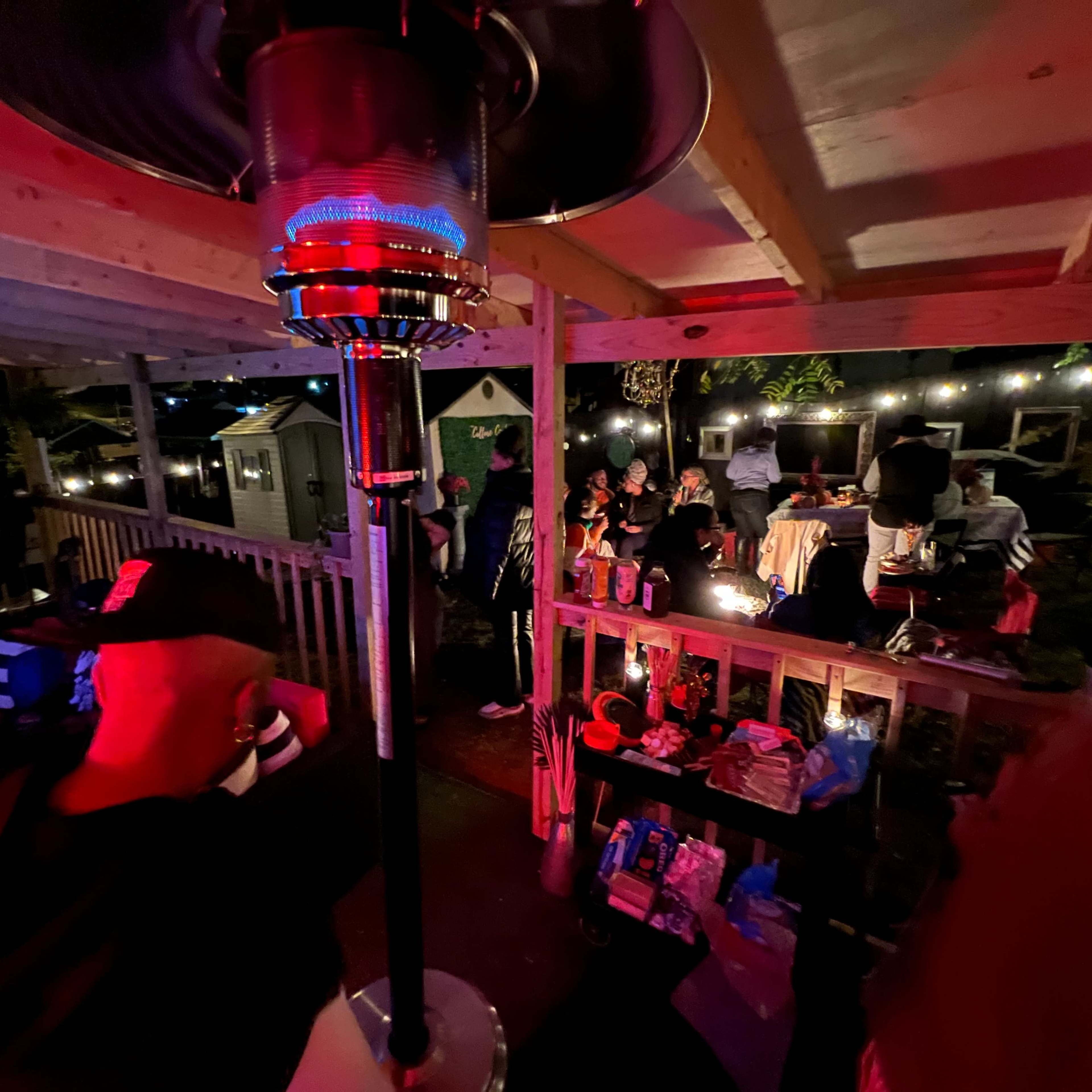 A gathering on a wooden patio, with people seated around tables and a patio heater illuminating the scene under string lights.