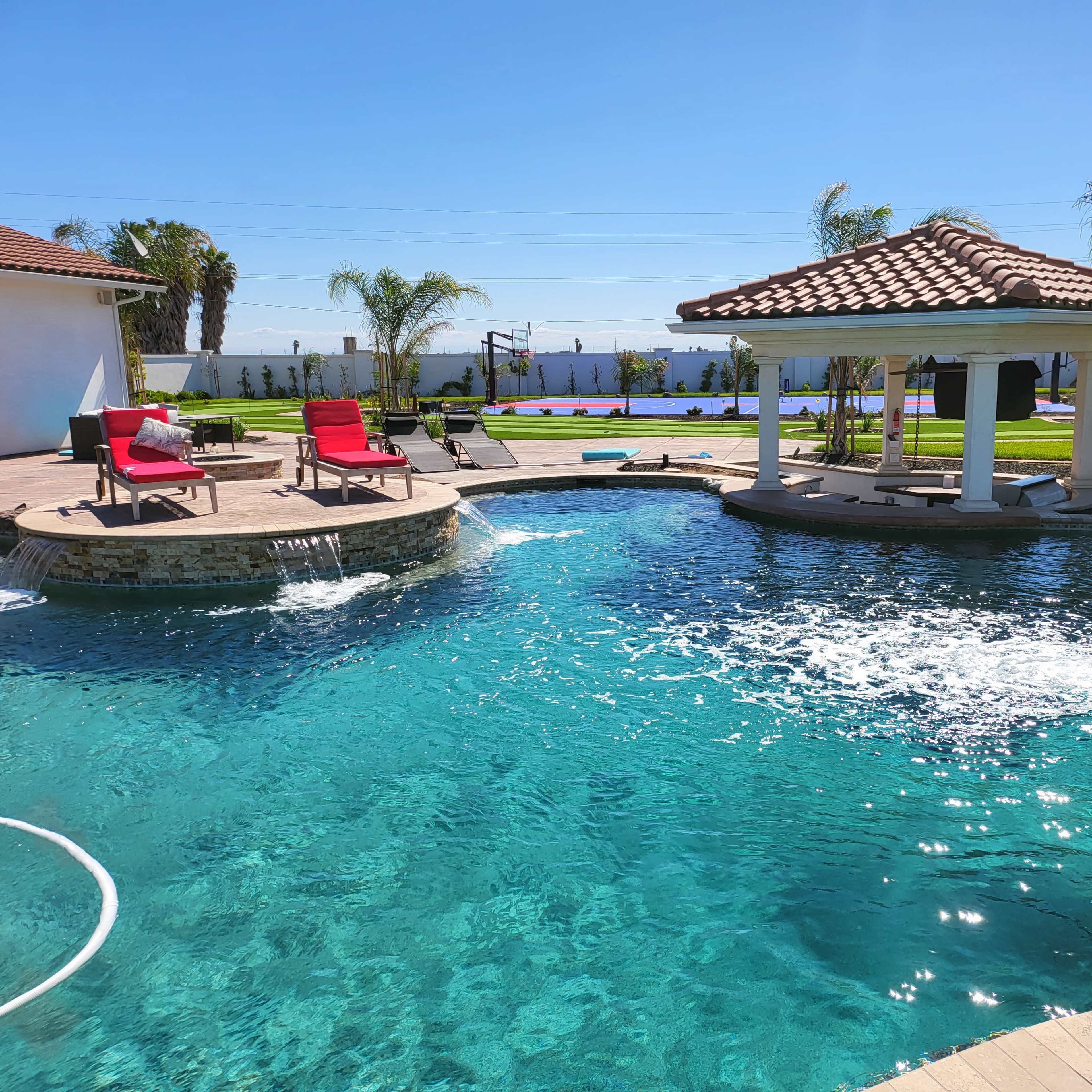A backyard with a swimming pool featuring a fountain, lounge chairs, and a gazebo surrounded by palm trees and landscaping.
