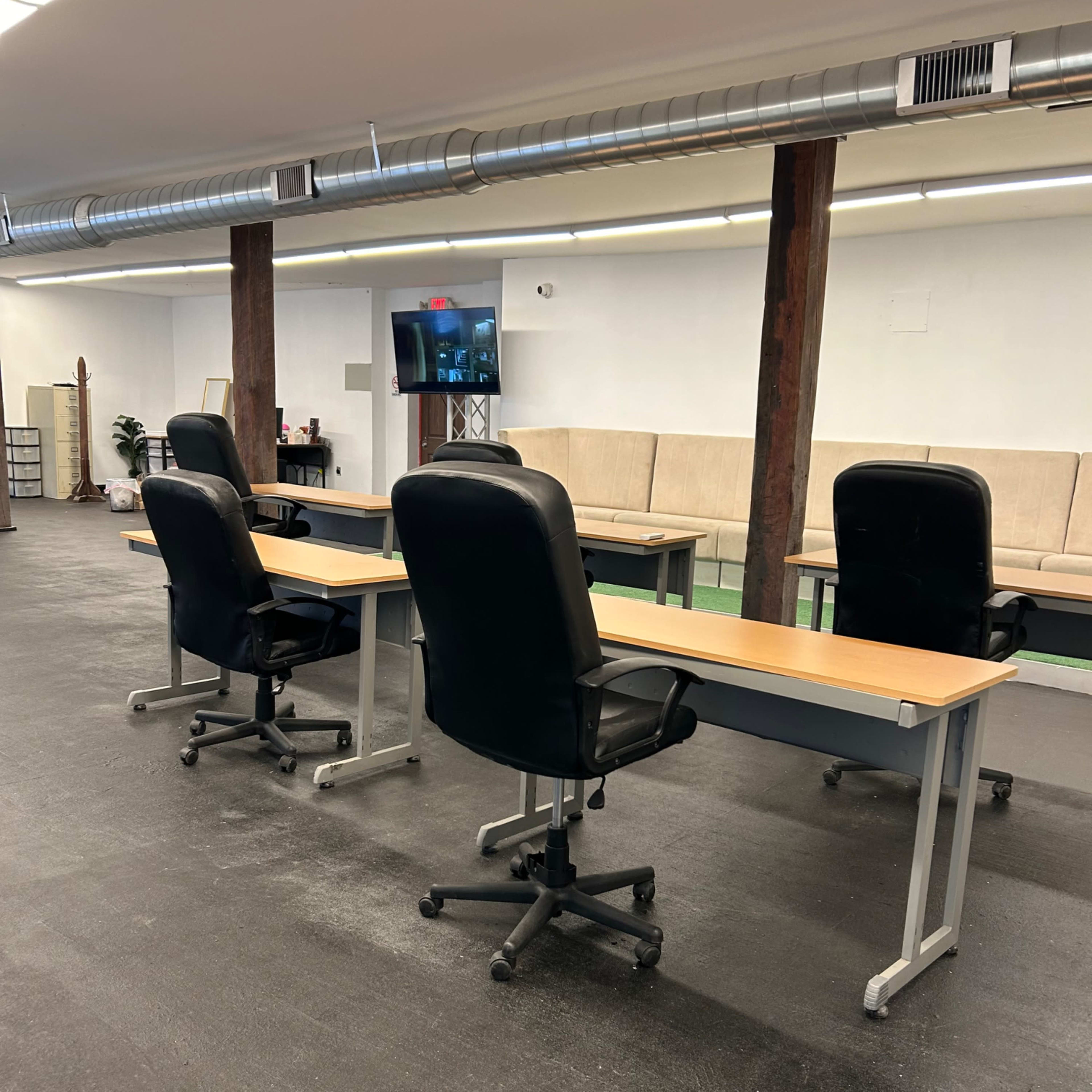A spacious office area with four black office chairs positioned at wooden desks, and a monitor visible in the background.