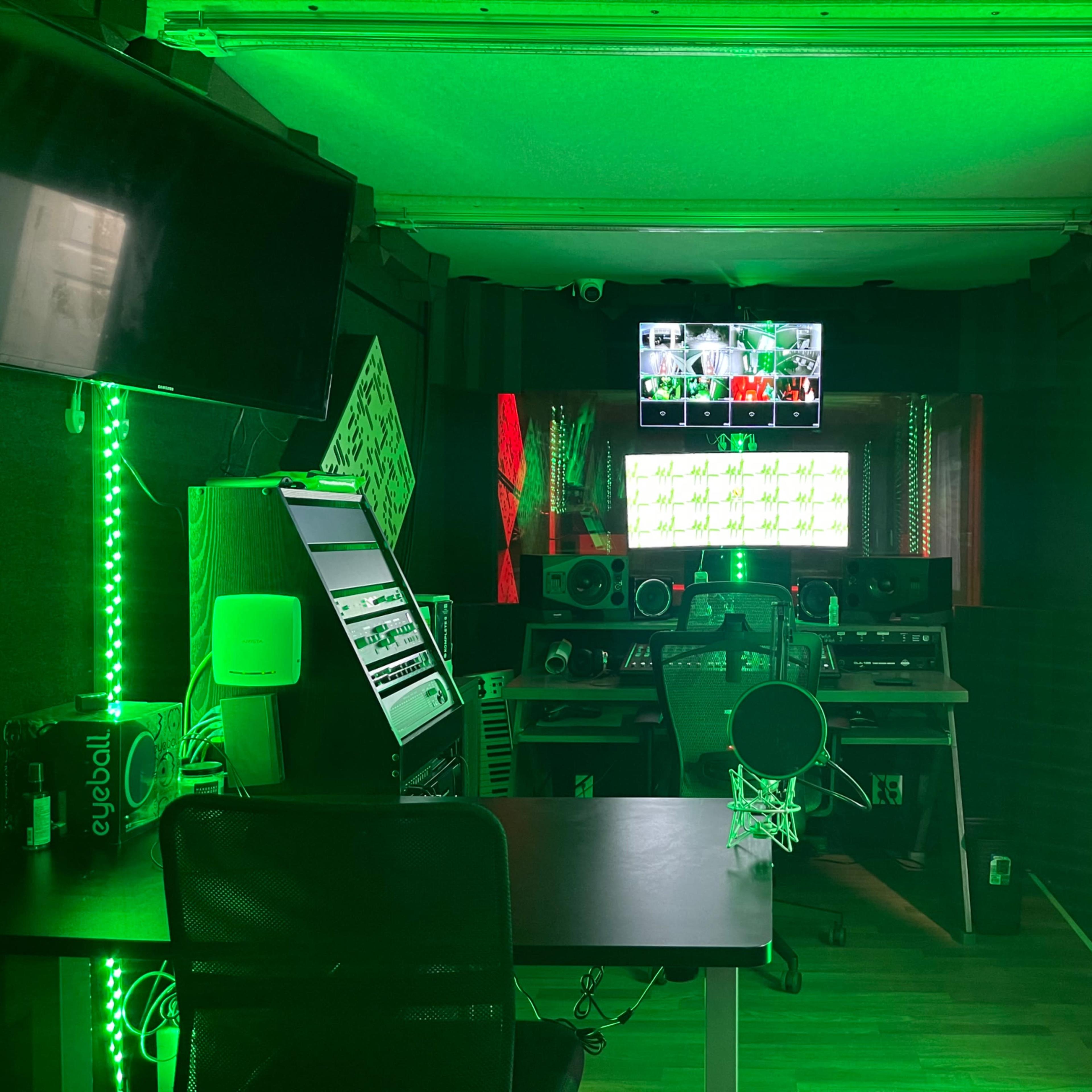 A music production studio featuring green LED lights, a large mixing console, and a computer monitor displaying visuals.