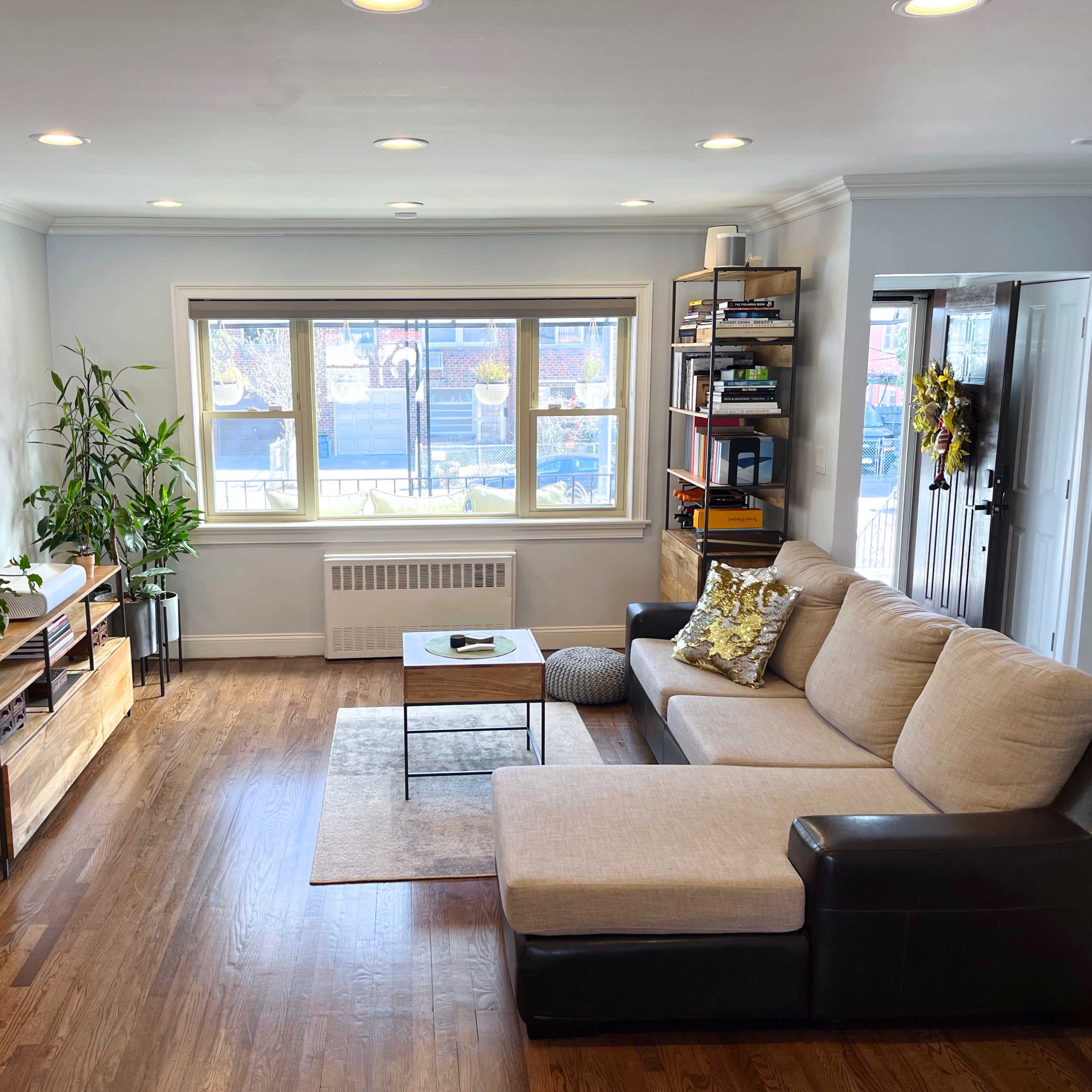 A modern living room with a light-colored sectional sofa, a wooden coffee table, a TV stand, and a large window allowing natural light to enter.
