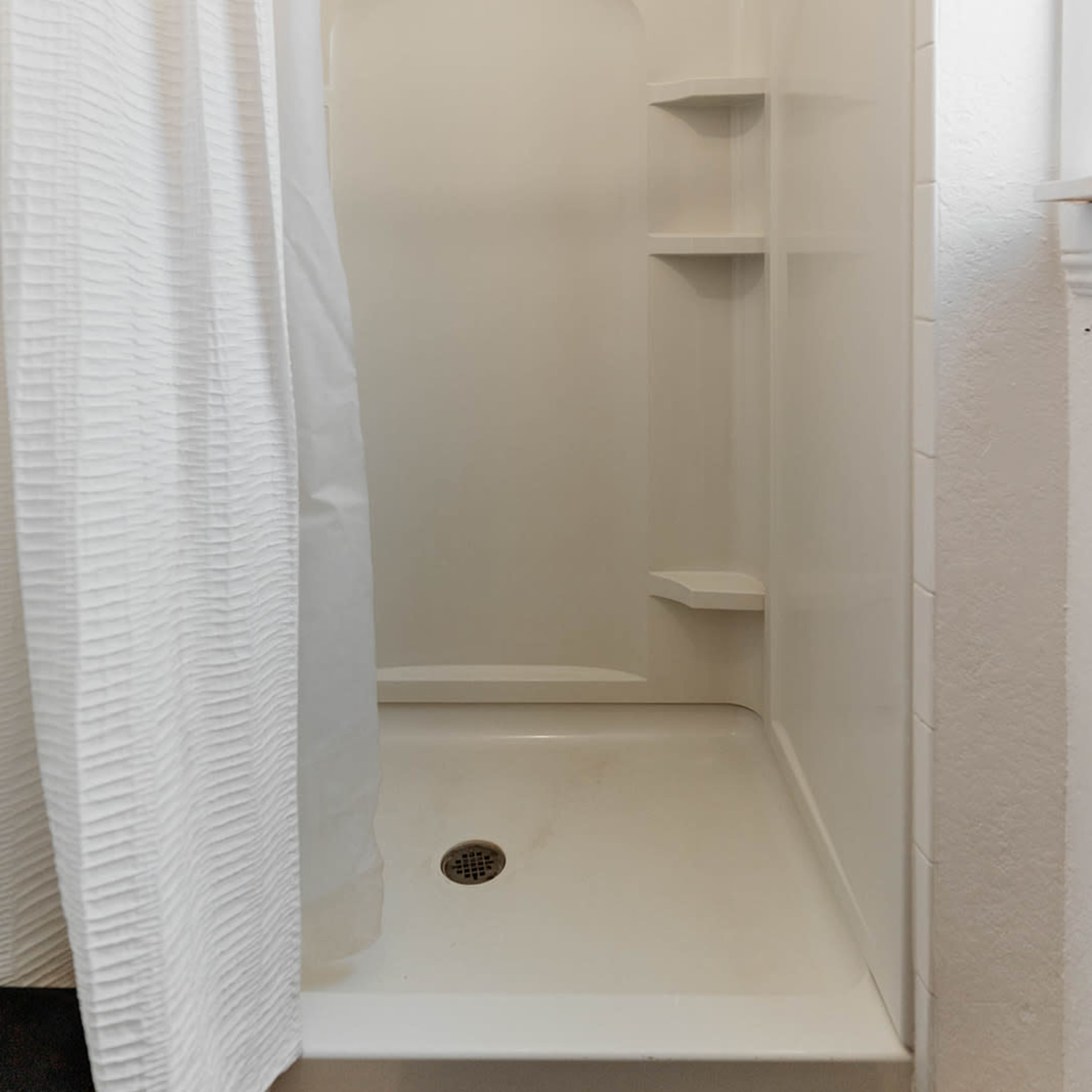 A clean, empty shower stall features white tiled walls, a showerhead, and a translucent curtain.