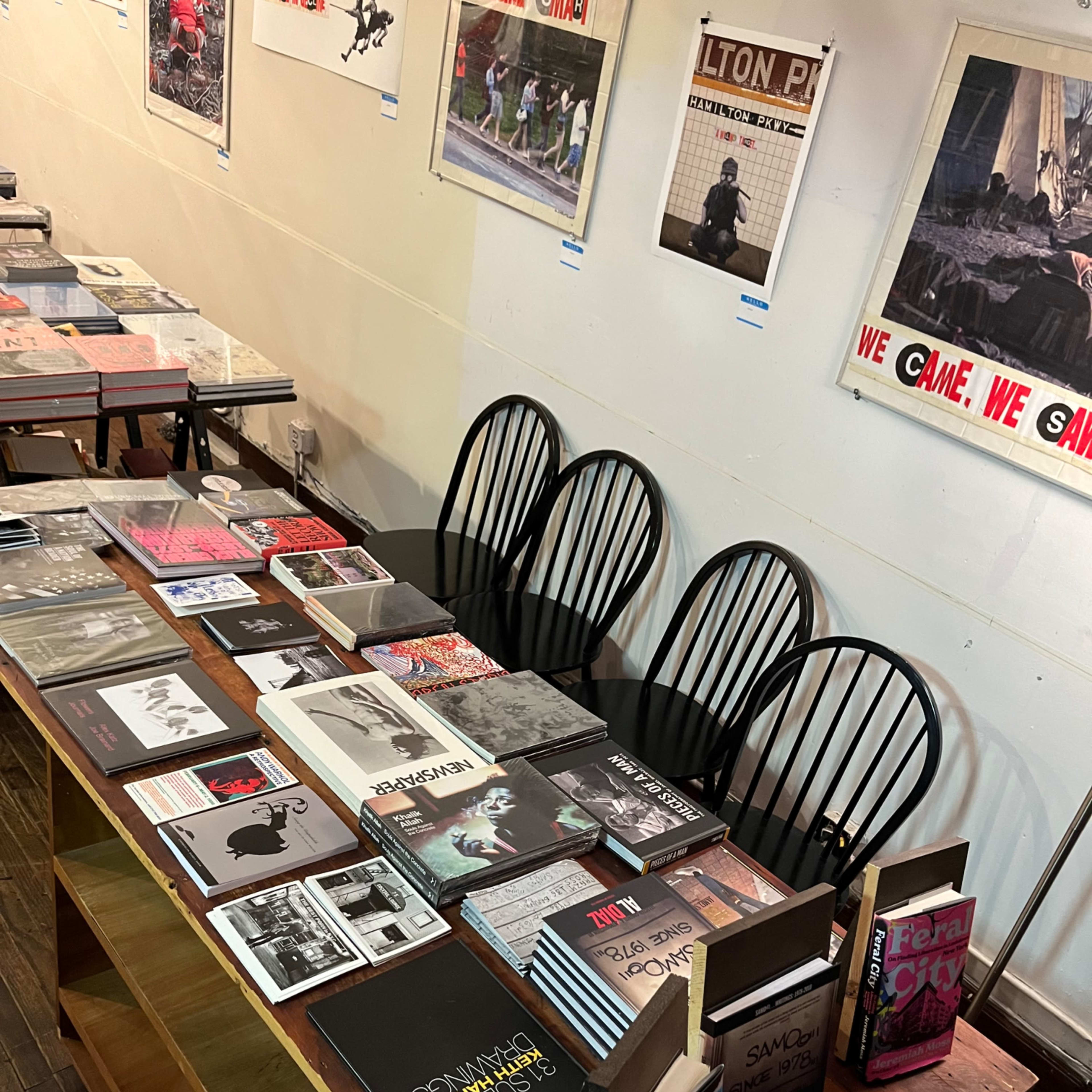 A long table displays various books and magazines, with several empty chairs arranged around it against a plain wall.