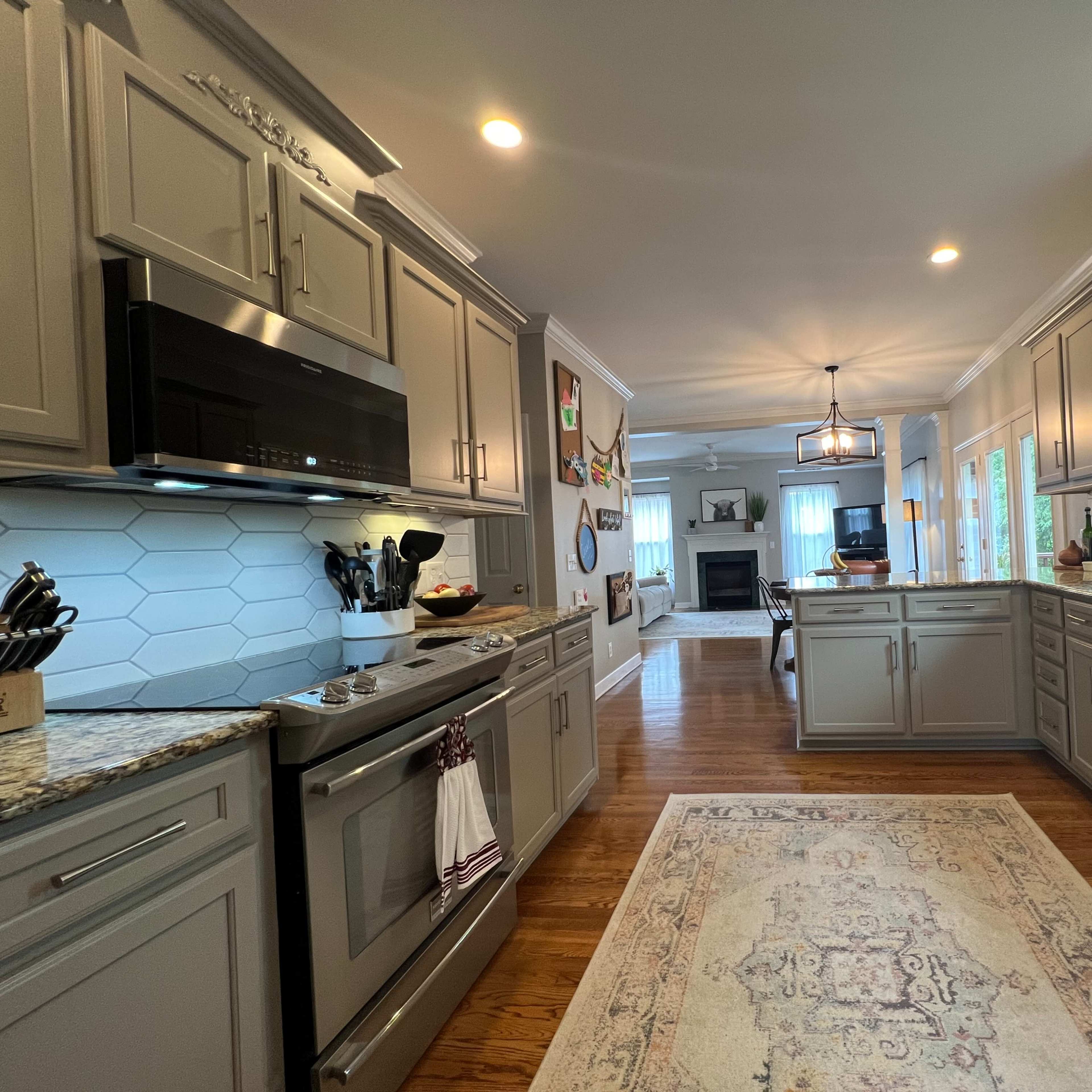 A spacious kitchen features gray cabinets, granite countertops, and a view into a living area with a fireplace.